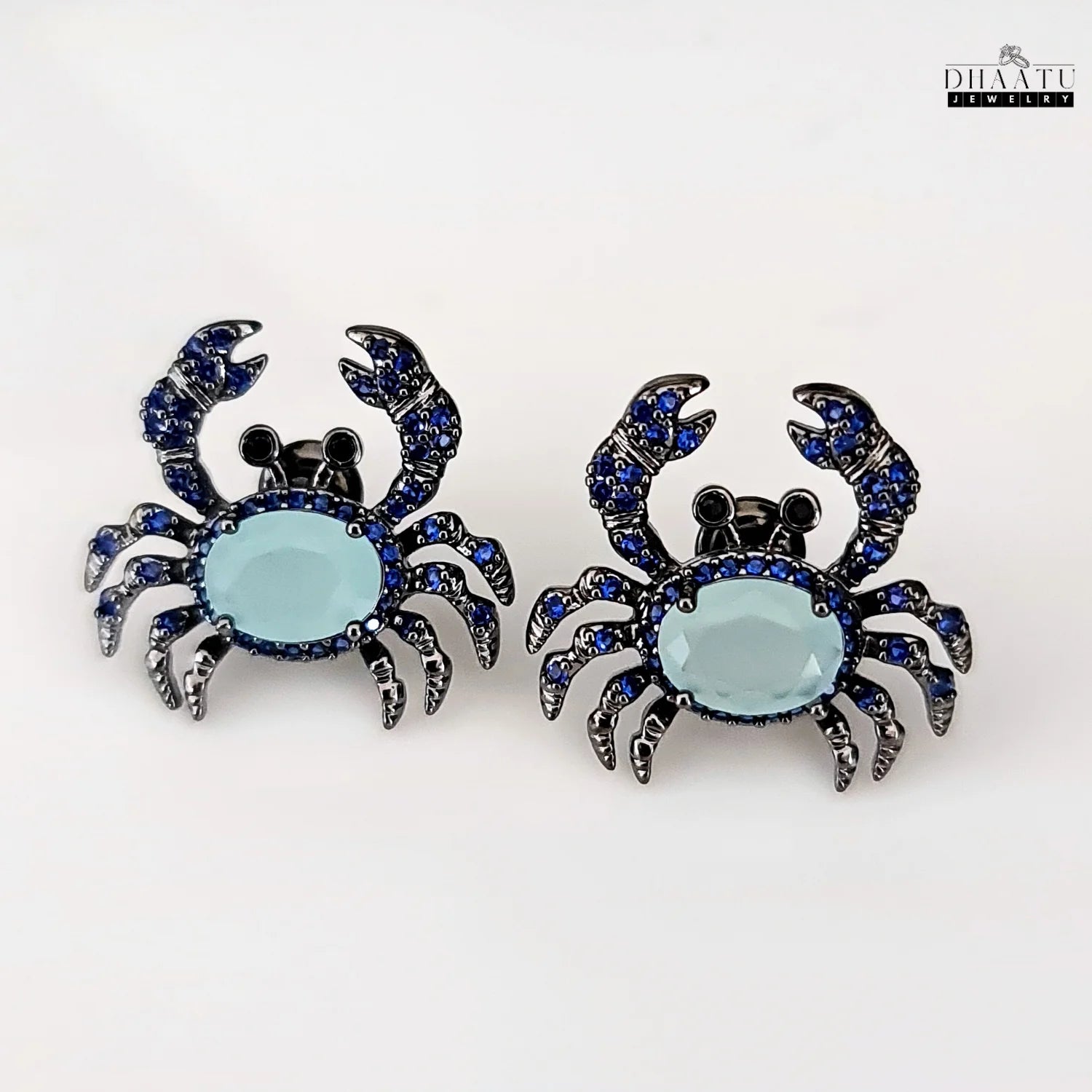Pair of silver-toned crab earrings with dark blue and aquamarine zircon stones on a white background.