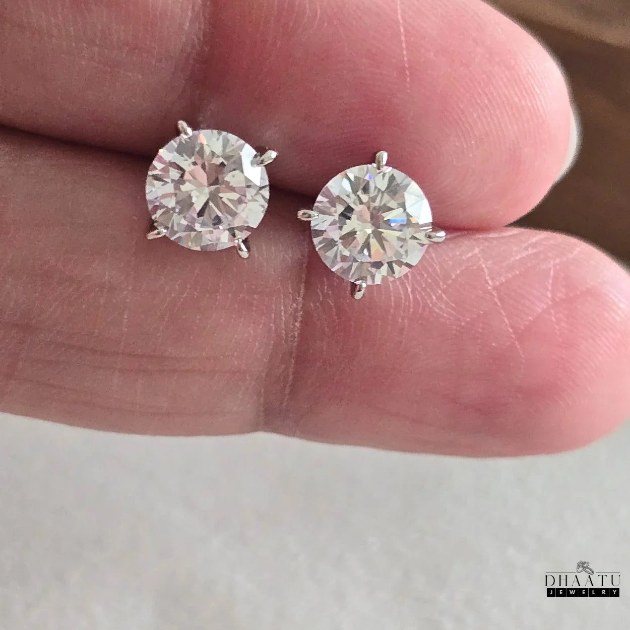 Two Dhaatu Jewelry silver-tone solitaire stud earrings, each with a sparkling cubic zirconia stone, held gently in a hand to show their size and brilliance.