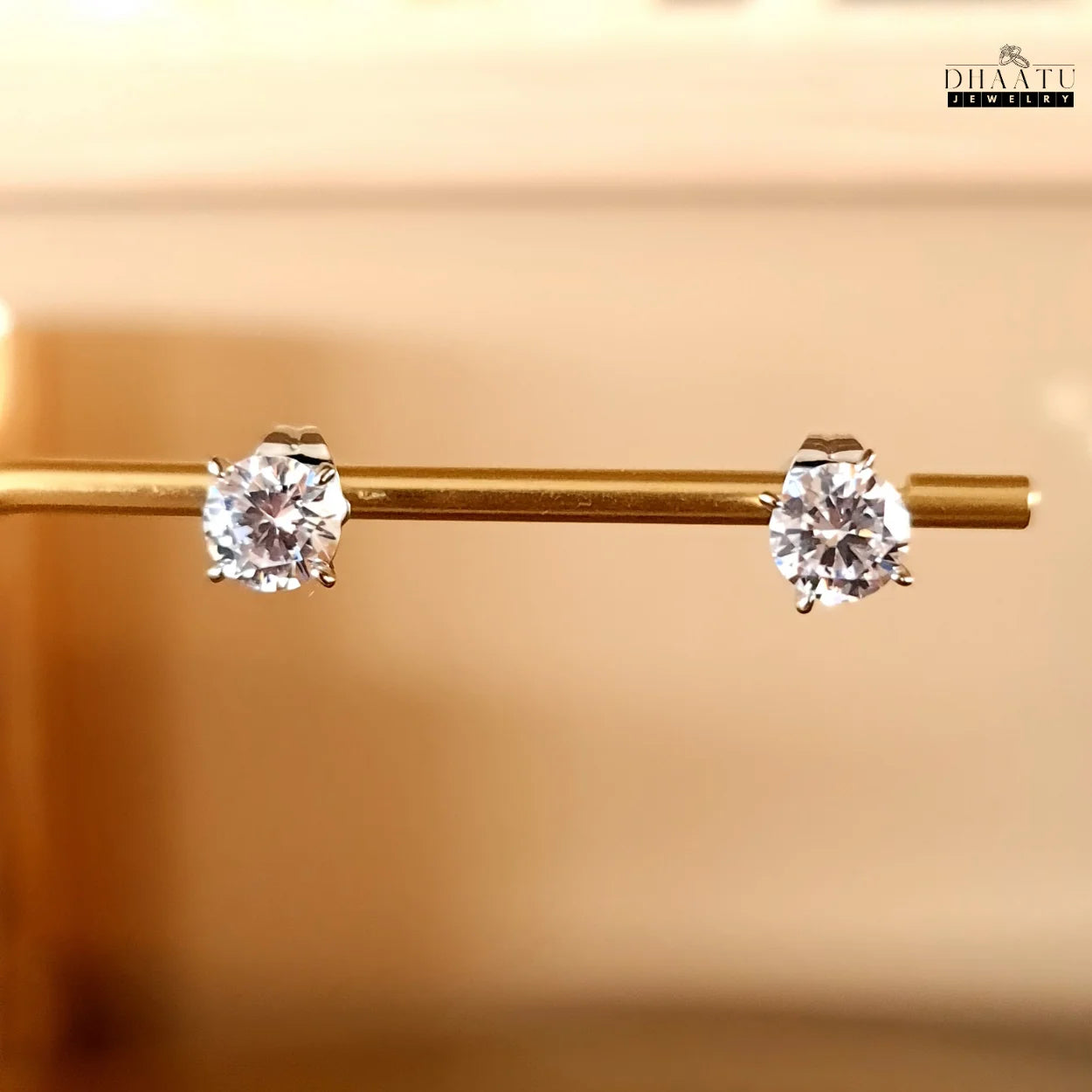 Dhaatu Jewelry silver-tone solitaire stud earrings with sparkling cubic zirconia gems, neatly arranged on a gold display bar, showcasing their perfect symmetry.