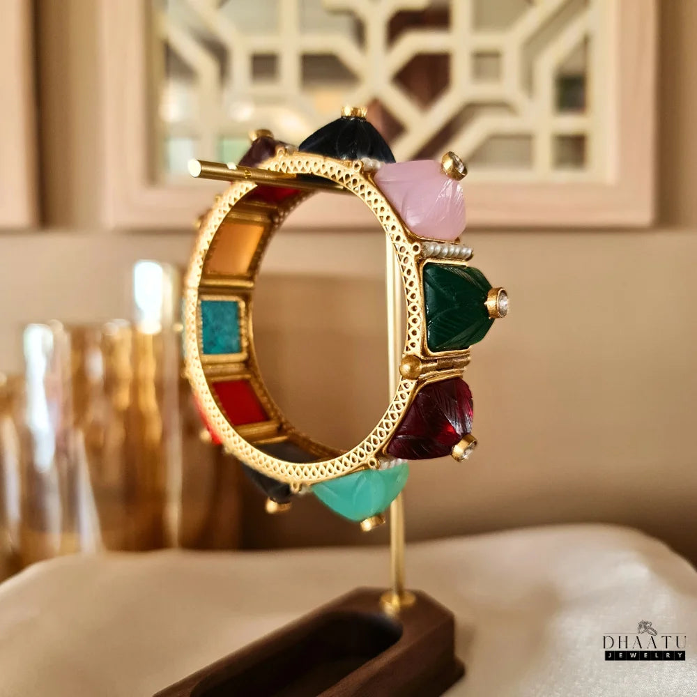 Side view of a multicolor carved stone and pearl kada bracelet on a clear glass stand, highlighting the full circular design with various colored stones and pearls.

