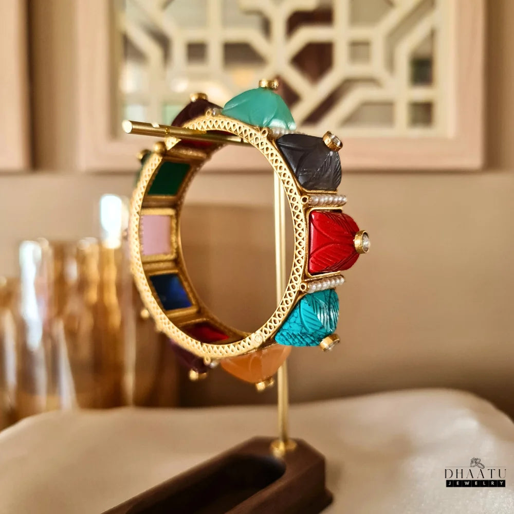 Close-up of a multicolor carved stone and pearl kada bracelet on a golden display stand, showing orange, maroon, black, and turquoise stones.

