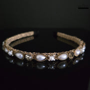 Close-up of elegant gold-tone hairband with white pearls and clear crystals on a black reflective surface, Dhaatu Jewelry modern design.