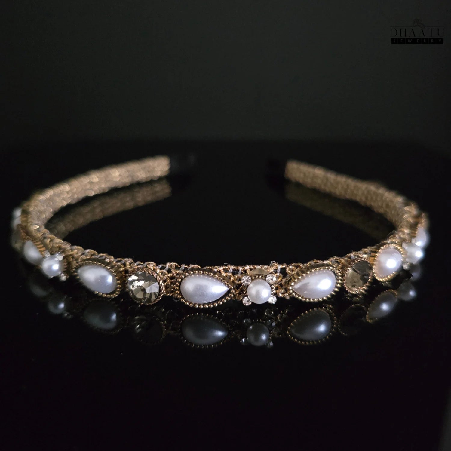 Close-up of elegant gold-tone hairband with white pearls and clear crystals on a black reflective surface, Dhaatu Jewelry modern design.