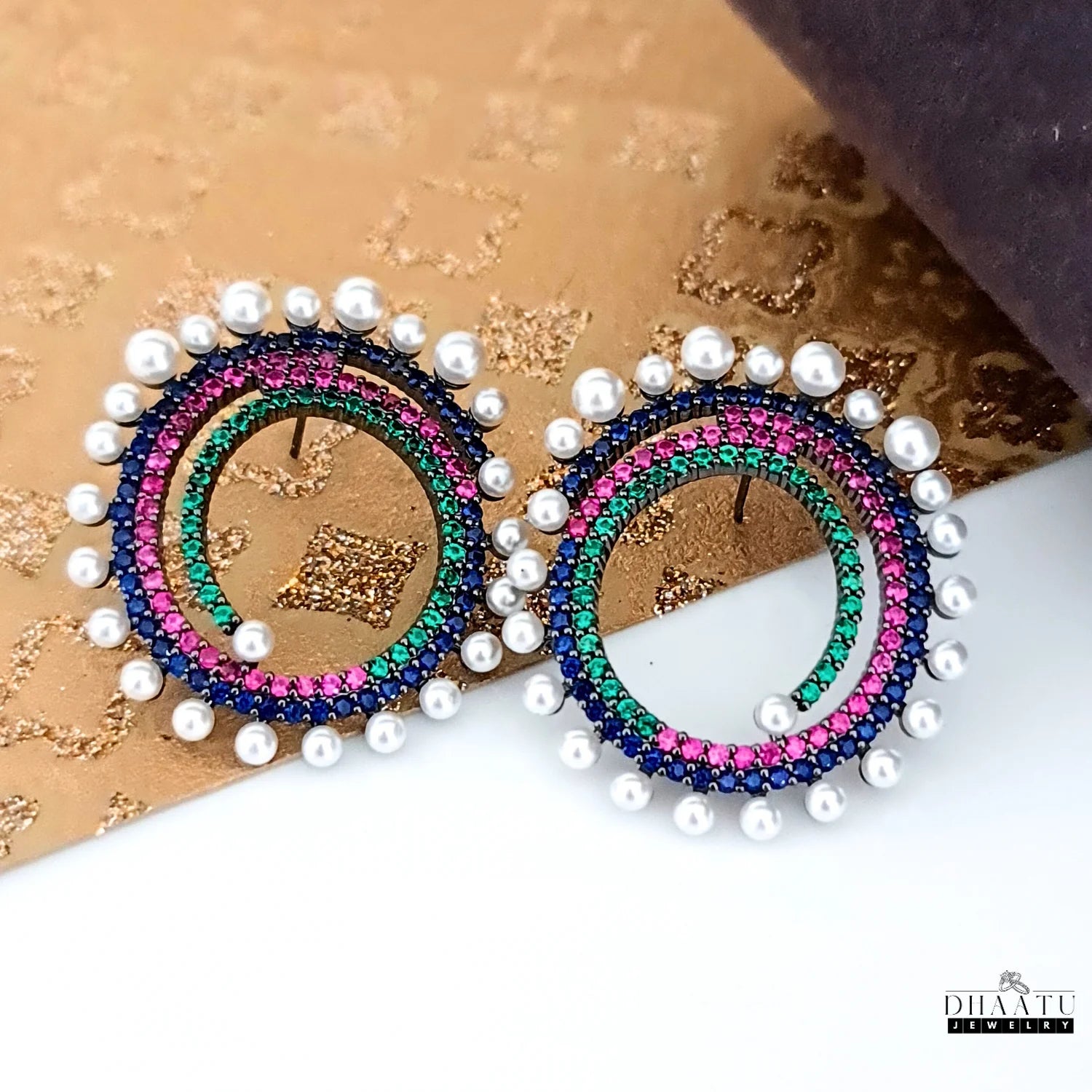 A pair of Dhaatu Jewelry festive designer earrings with spiral gemstone and pearl design, placed on a decorative gold and brown textured background.