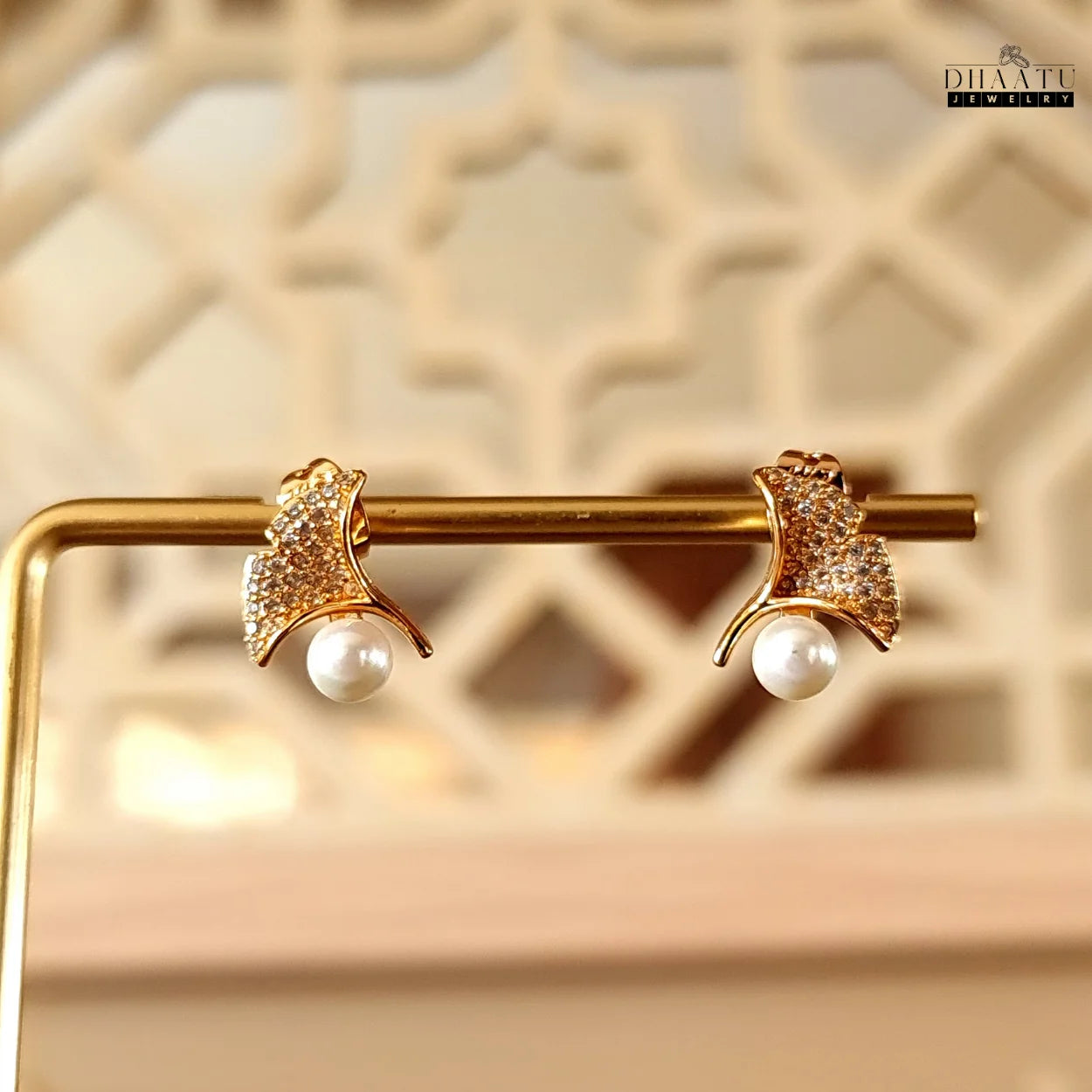 Gold ginkgo leaf stud earrings with faux pearls and cubic zirconia on a gold display stand with a decorative background
