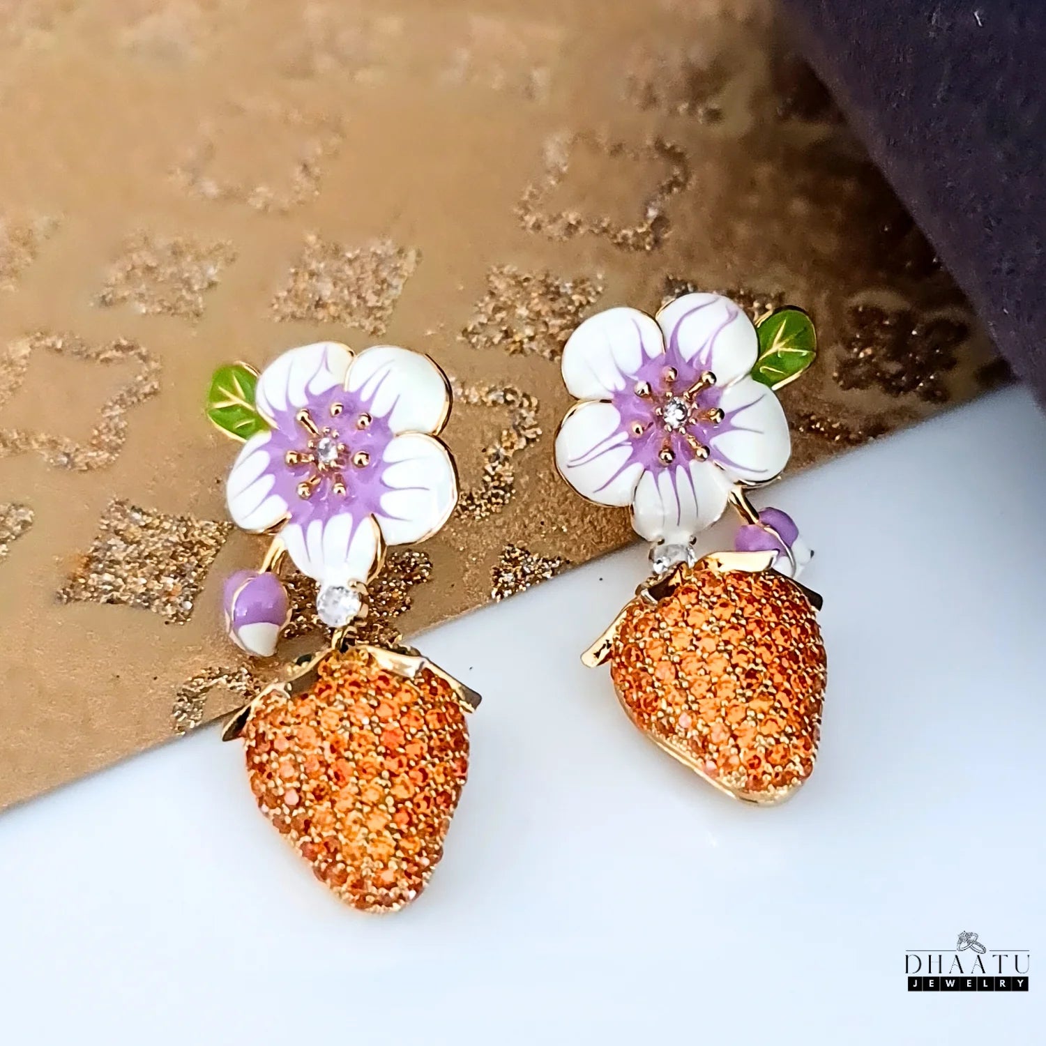 Gold-plated drop earrings with white and purple enamel flowers and orange CZ strawberry charms, displayed on a textured gold and white surface.