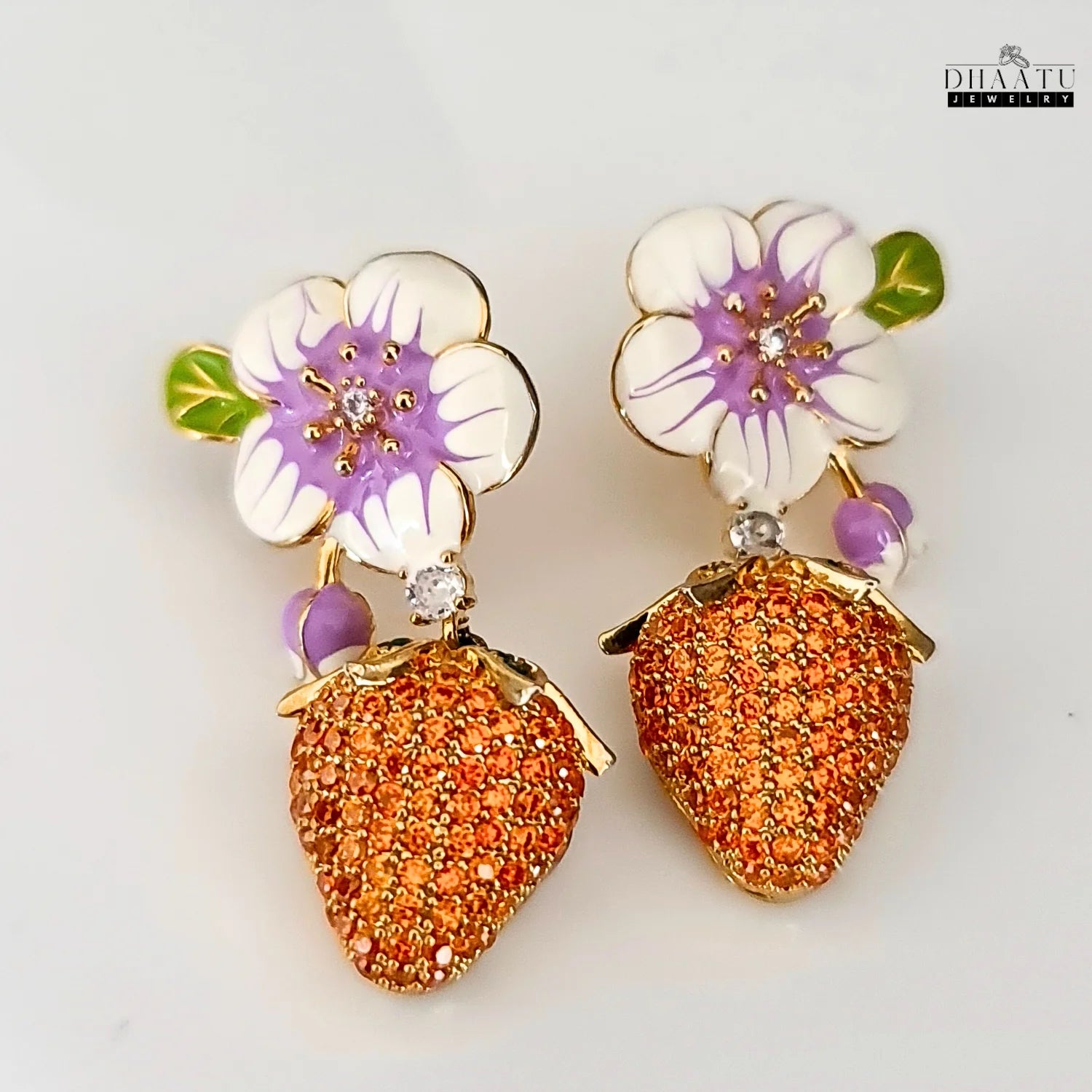Pair of gold-plated drop earrings featuring white and purple enamel flowers with orange CZ strawberry charms, displayed on a white background.