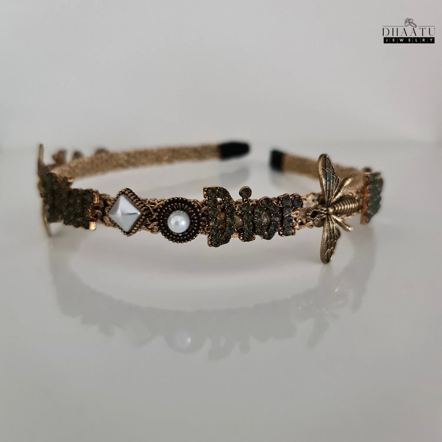 Front view of golden hairband with bee, "Dior" rhinestones, and pearls on a white background