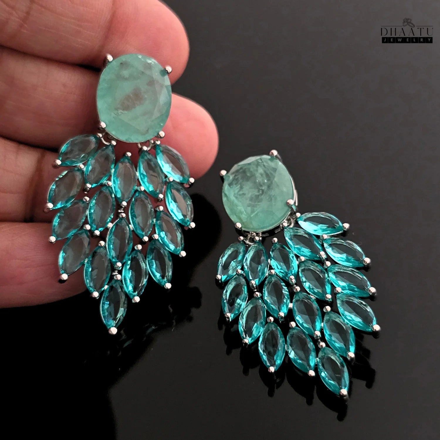 A hand holding one of the Aqua Chalcedony and marquise cut blue stone dangler earrings, providing a sense of scale and detail against a dark background.