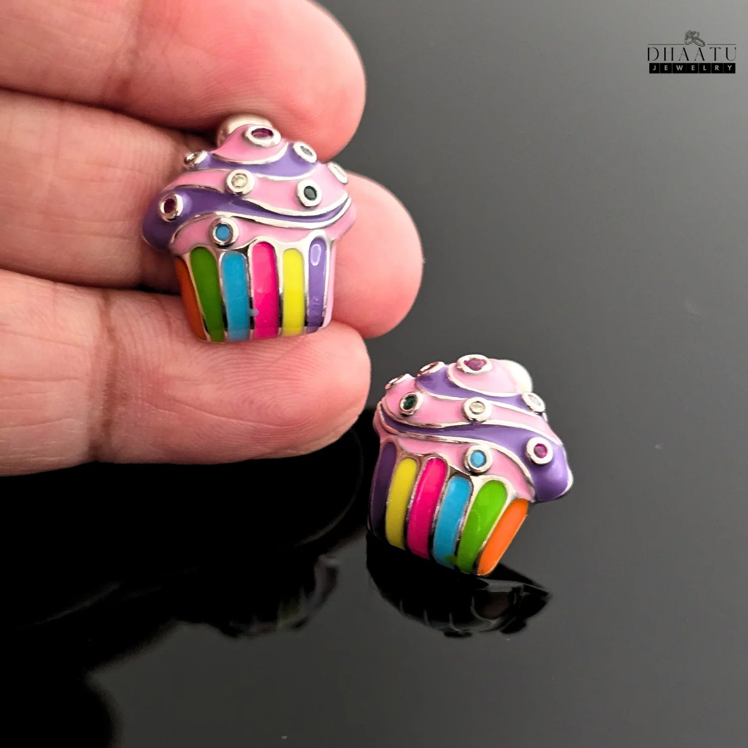 A hand holding one of the colorful cupcake enamel earrings, providing a sense of scale and detail against a black reflective background.