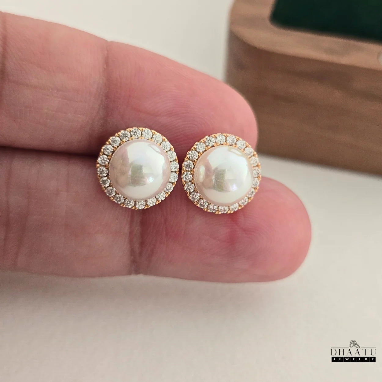A hand holding up two round pearl and zircon halo stud earrings, showcasing their details.