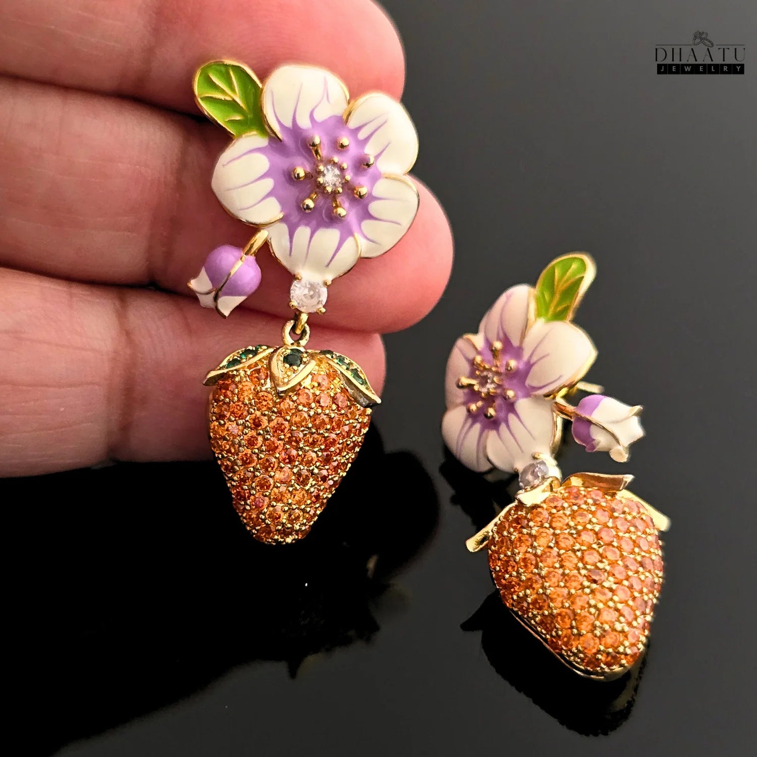 Close-up of a hand holding a gold-plated drop earring with a white and purple enamel flower and an orange CZ strawberry charm, on a black background.