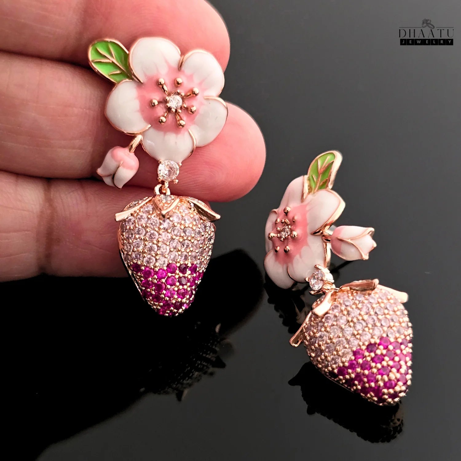 Delicate earrings with floral and berry designs held by a hand on a dark background.