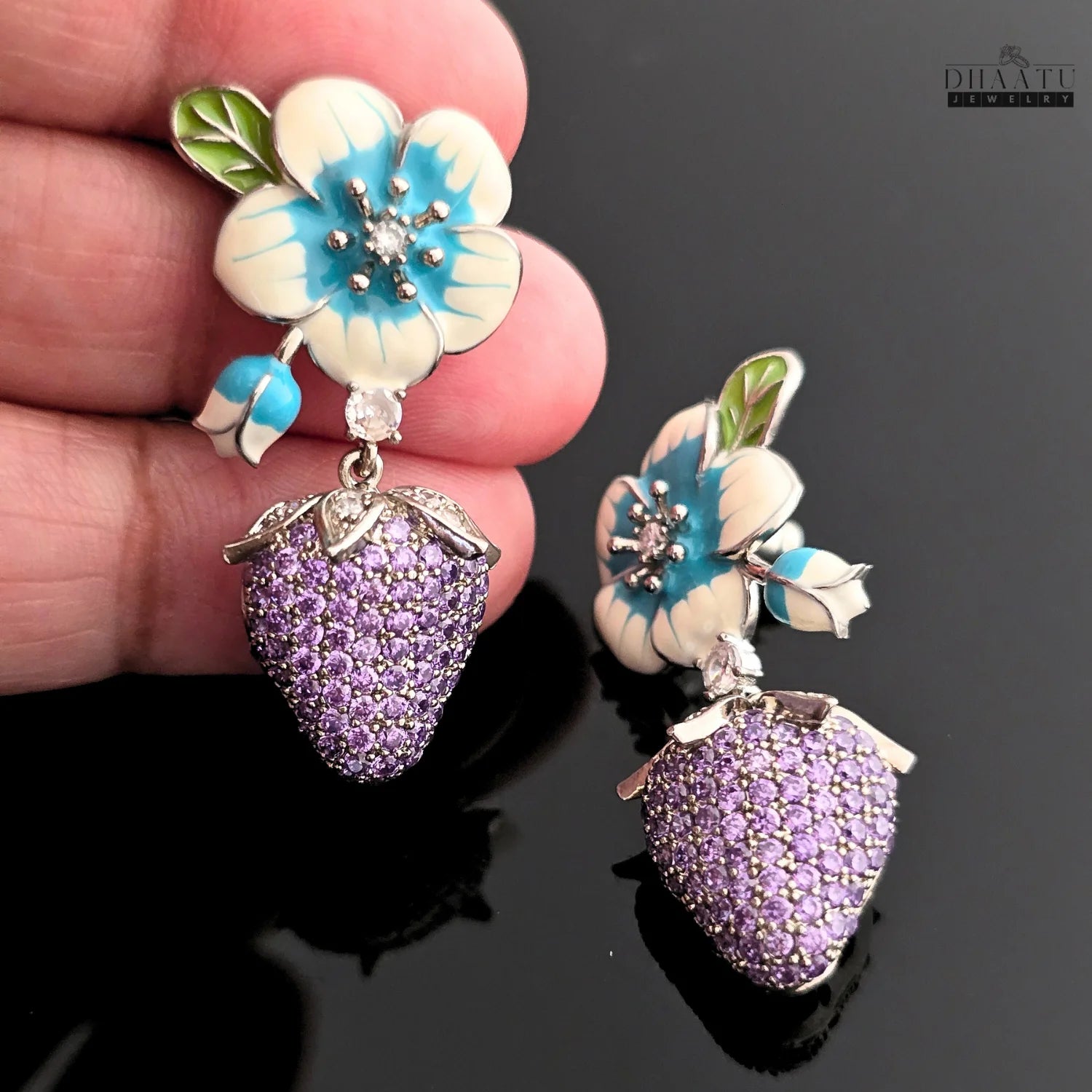 Close-up of a hand holding a silver-plated drop earring with a white and blue enamel flower and a purple CZ strawberry charm, on a black background.