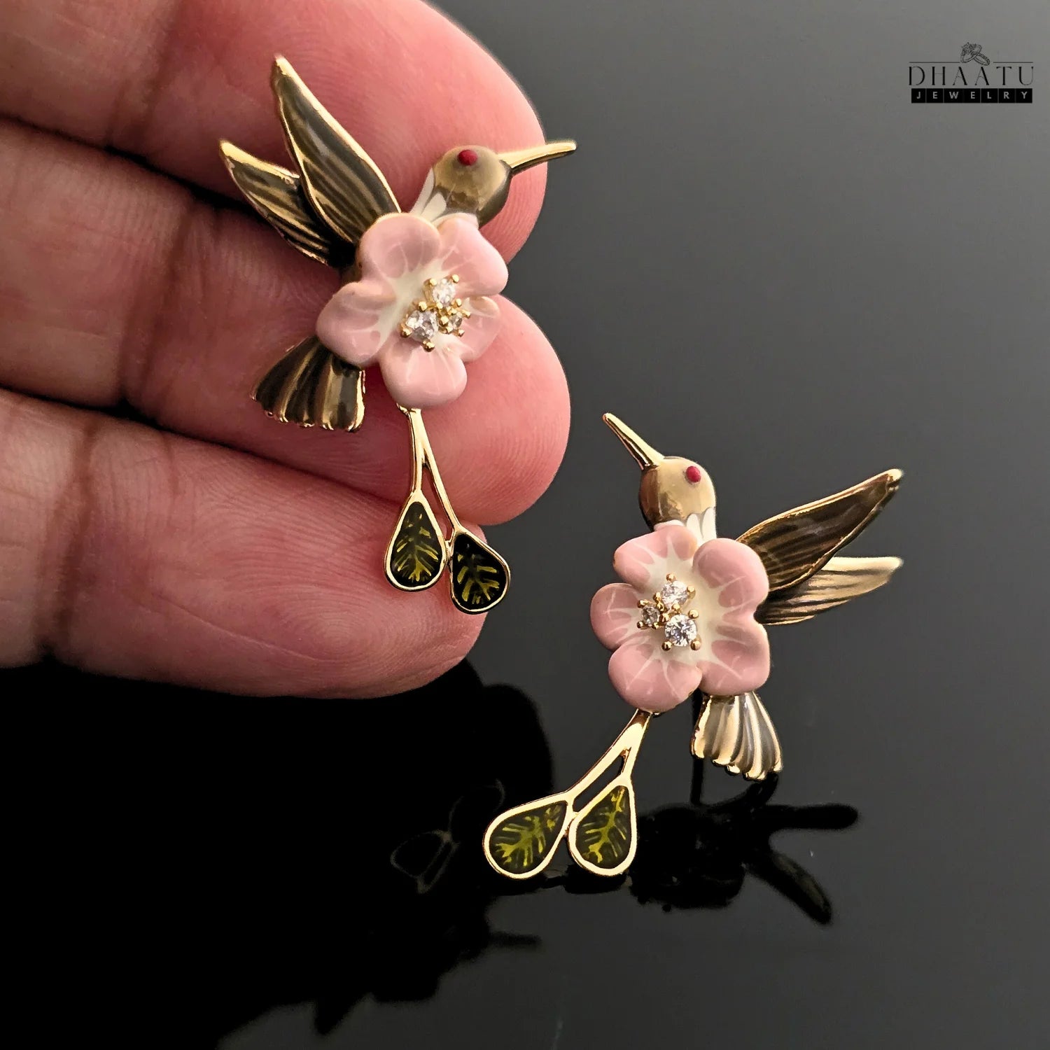 Close-up shot of a pink and brown enamel hummingbird earring being held, showing the detailed pink flower and hanging leaves.