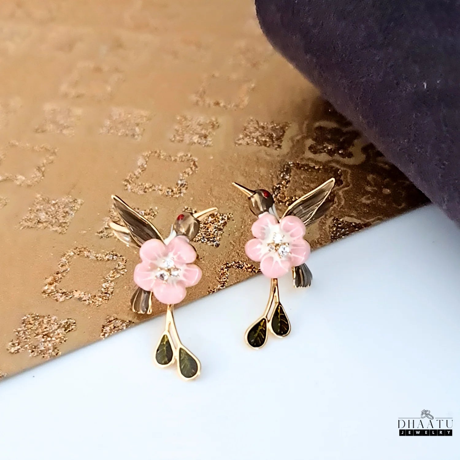 A pair of pink and brown enamel hummingbird earrings styled on a textured gold and purple background.