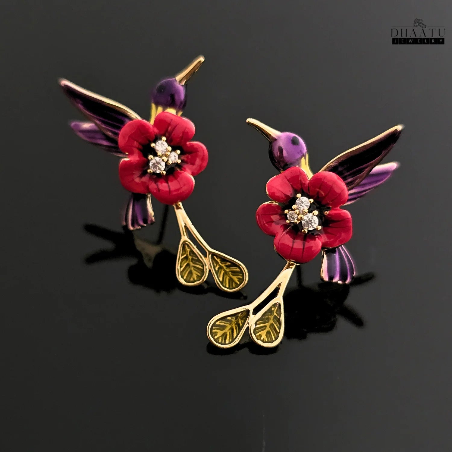 A pair of handcrafted red and purple enamel hummingbird and flower drop earrings on a black reflective surface.