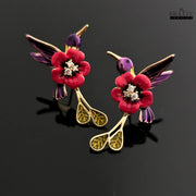 A pair of handcrafted red and purple enamel hummingbird and flower drop earrings on a black reflective surface.