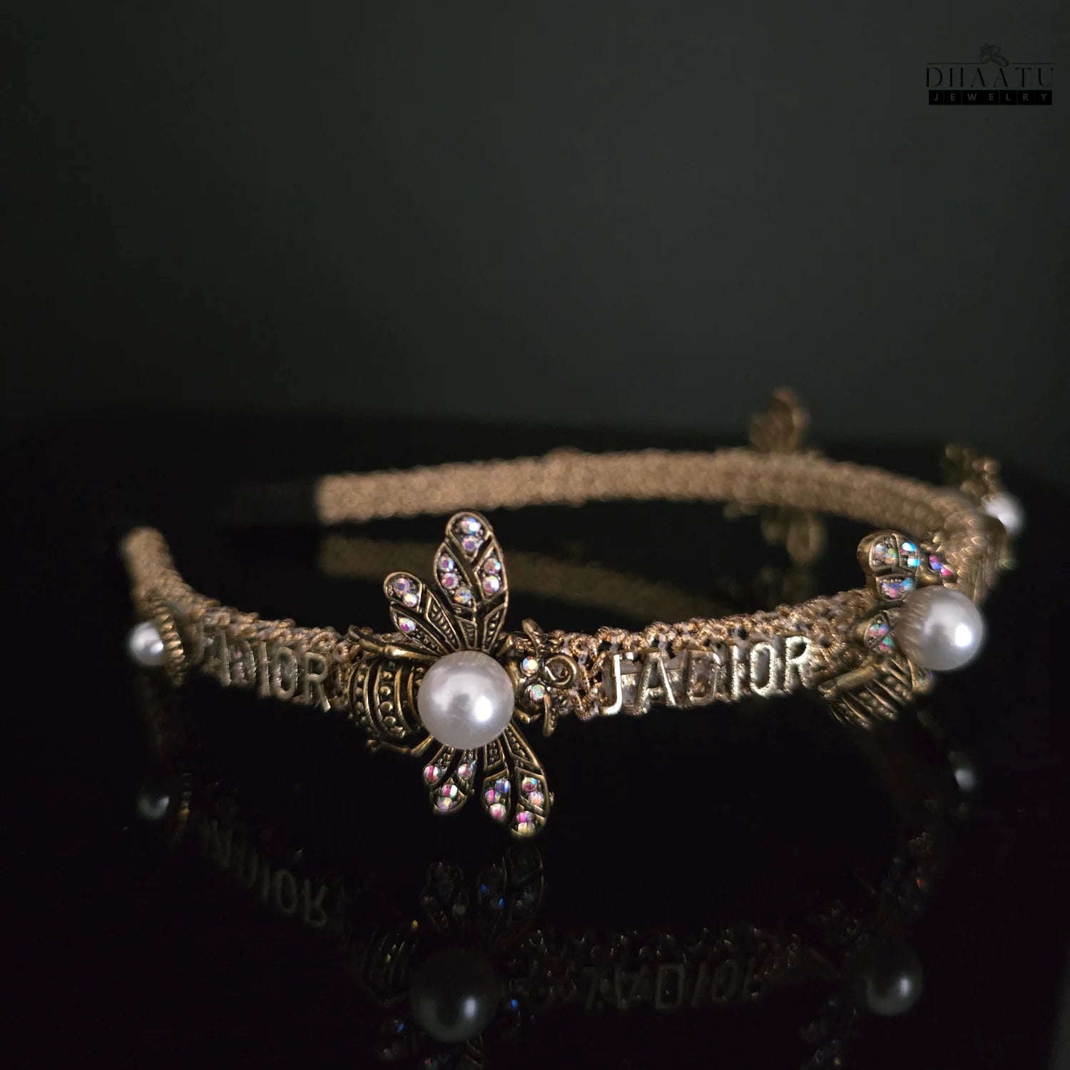 Side view of a modern golden hairband featuring detailed bee designs, iridescent crystals, and white pearls on a reflective dark surface