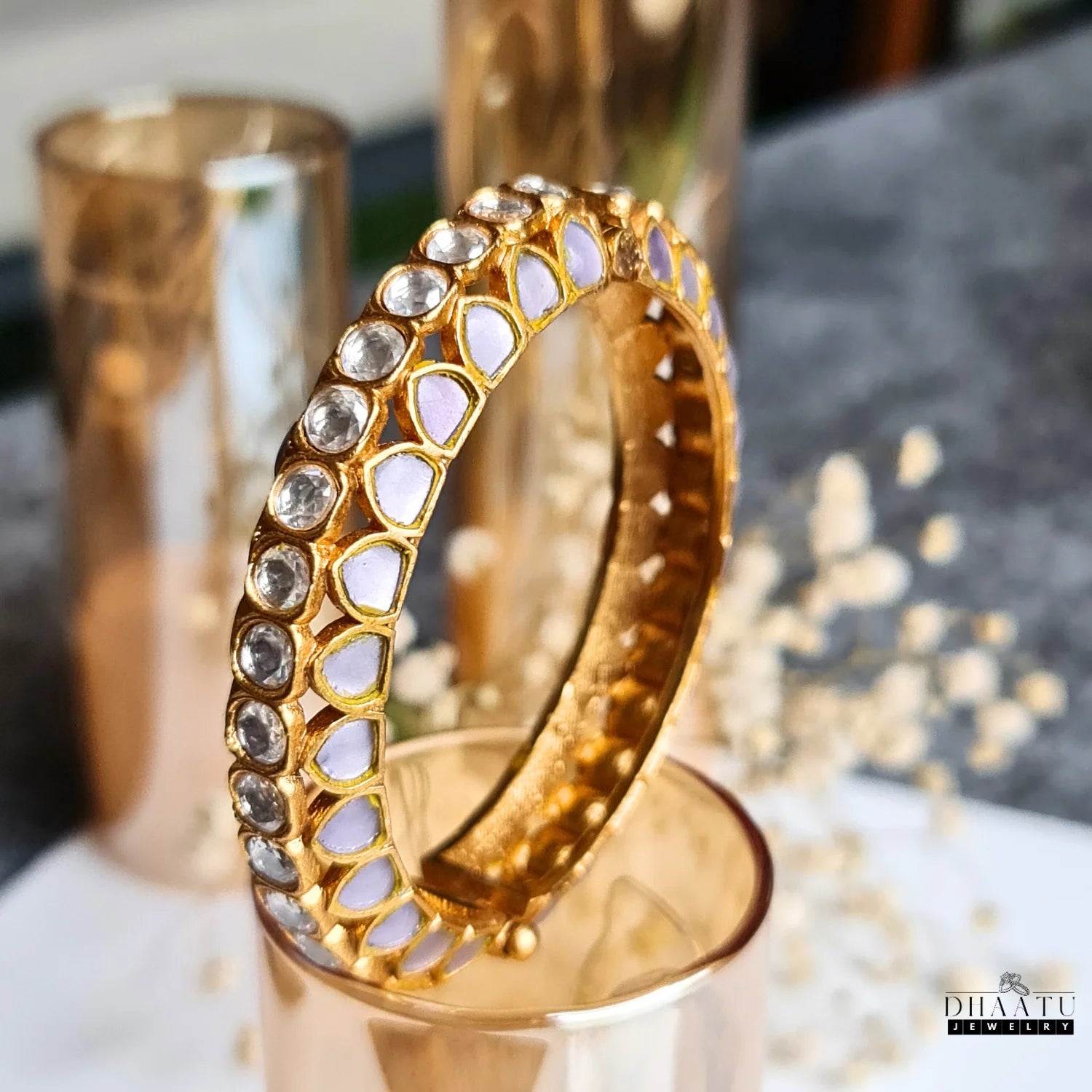 Gold-plated Jadau and Kemp stone kada in lavender, featuring clear and purple stones, displayed elegantly on a gold-colored glass, with a blurred background.

