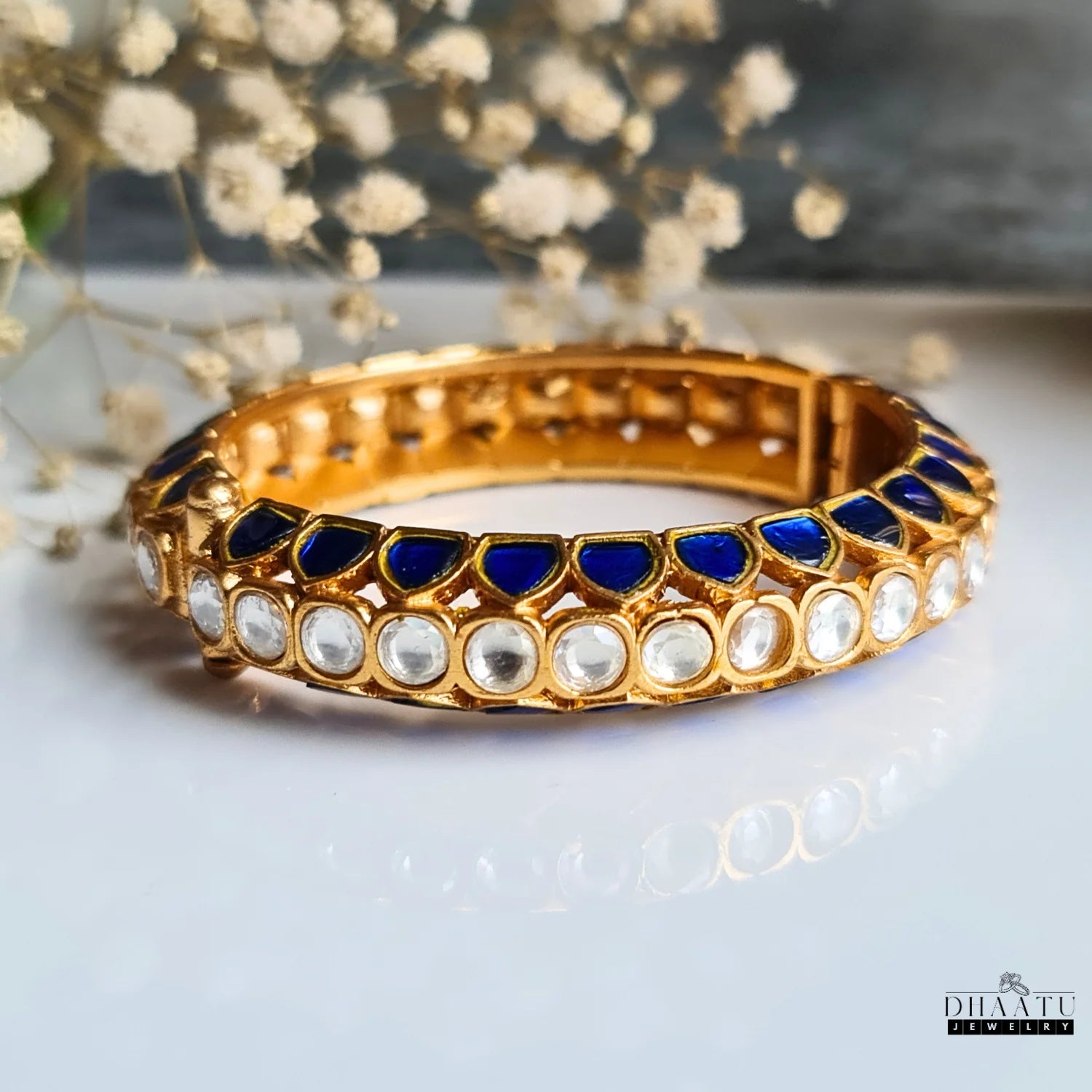 Gold-plated Jadau and Kemp stone kada in sapphire blue with clear and blue stones, viewed from an angle showing its reflection on a white surface with a subtle floral background.

