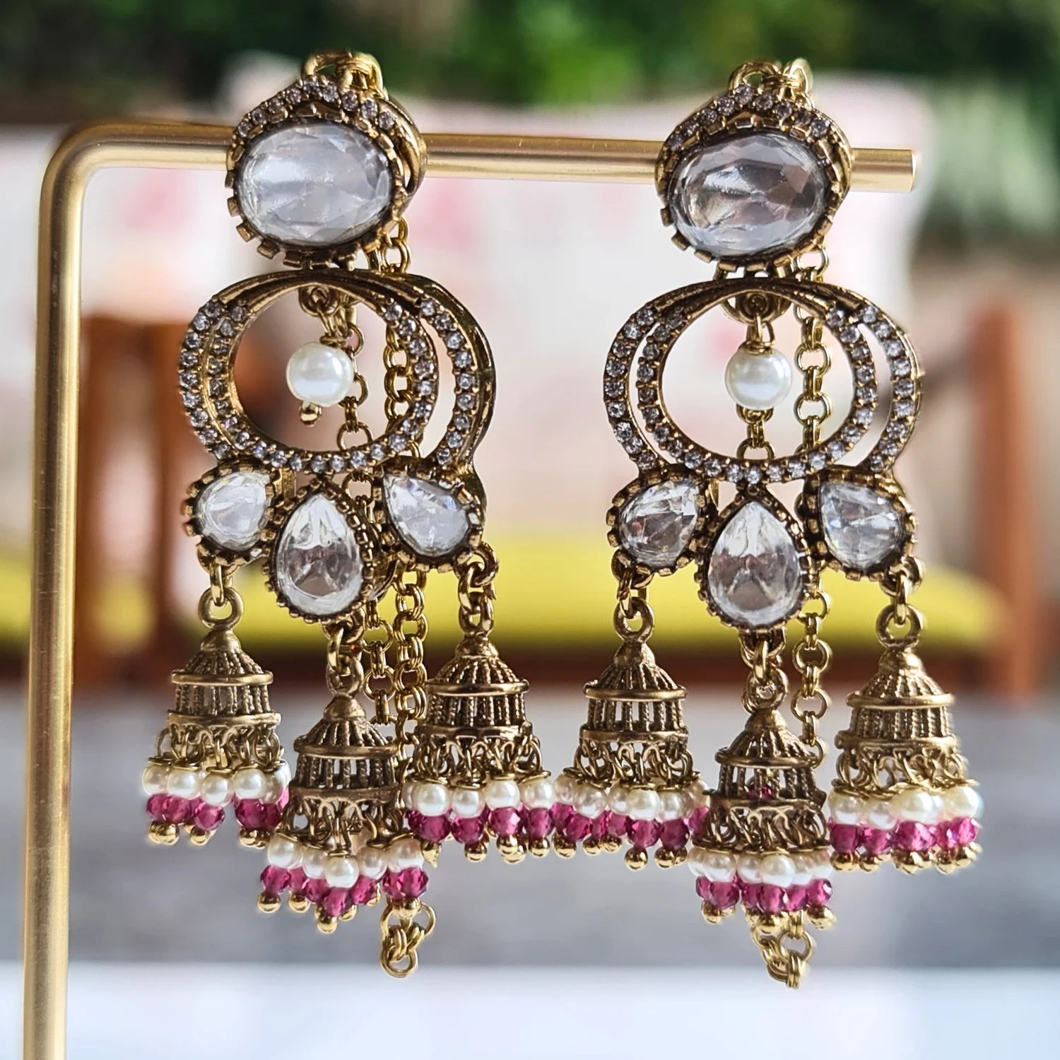 Antique gold-tone Kundan jhumka earrings with maroon pearl drops, showcased on a golden jewelry stand with outdoor lighting.

