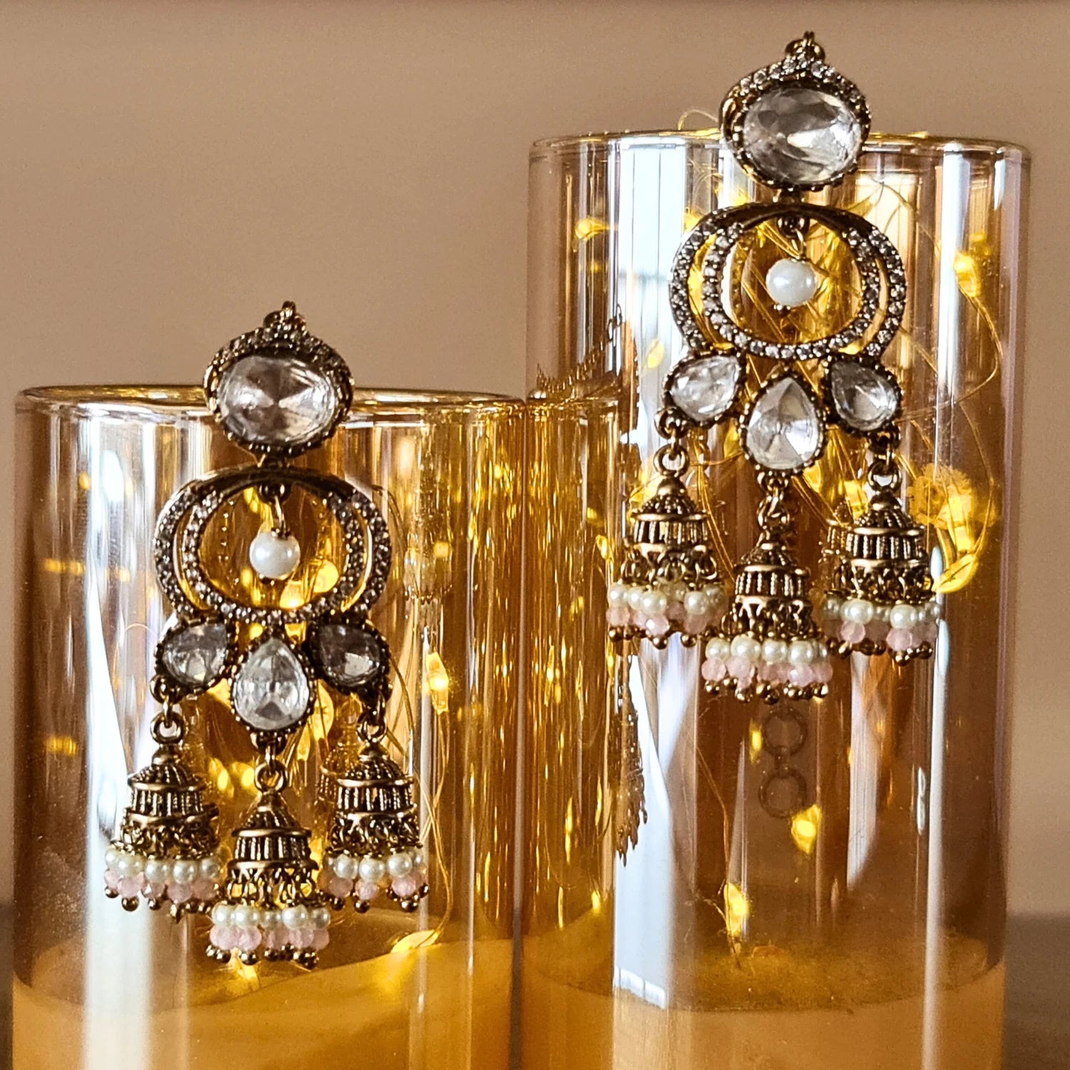 Beautiful Kundan Jhumka earrings with light pink pearl beads, elegantly placed on glowing golden glass cylinders.

