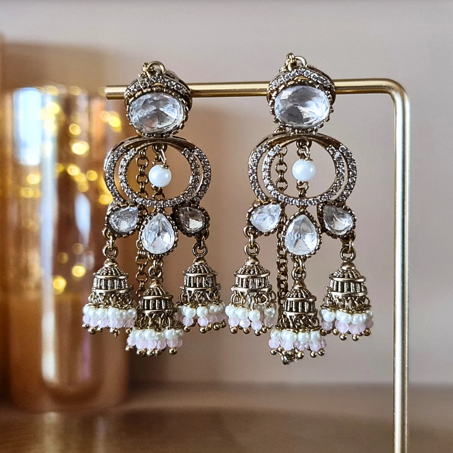 Antique gold-tone Kundan jhumka earrings with light pink pearl drops, showcased on a golden jewelry stand with warm lighting.



