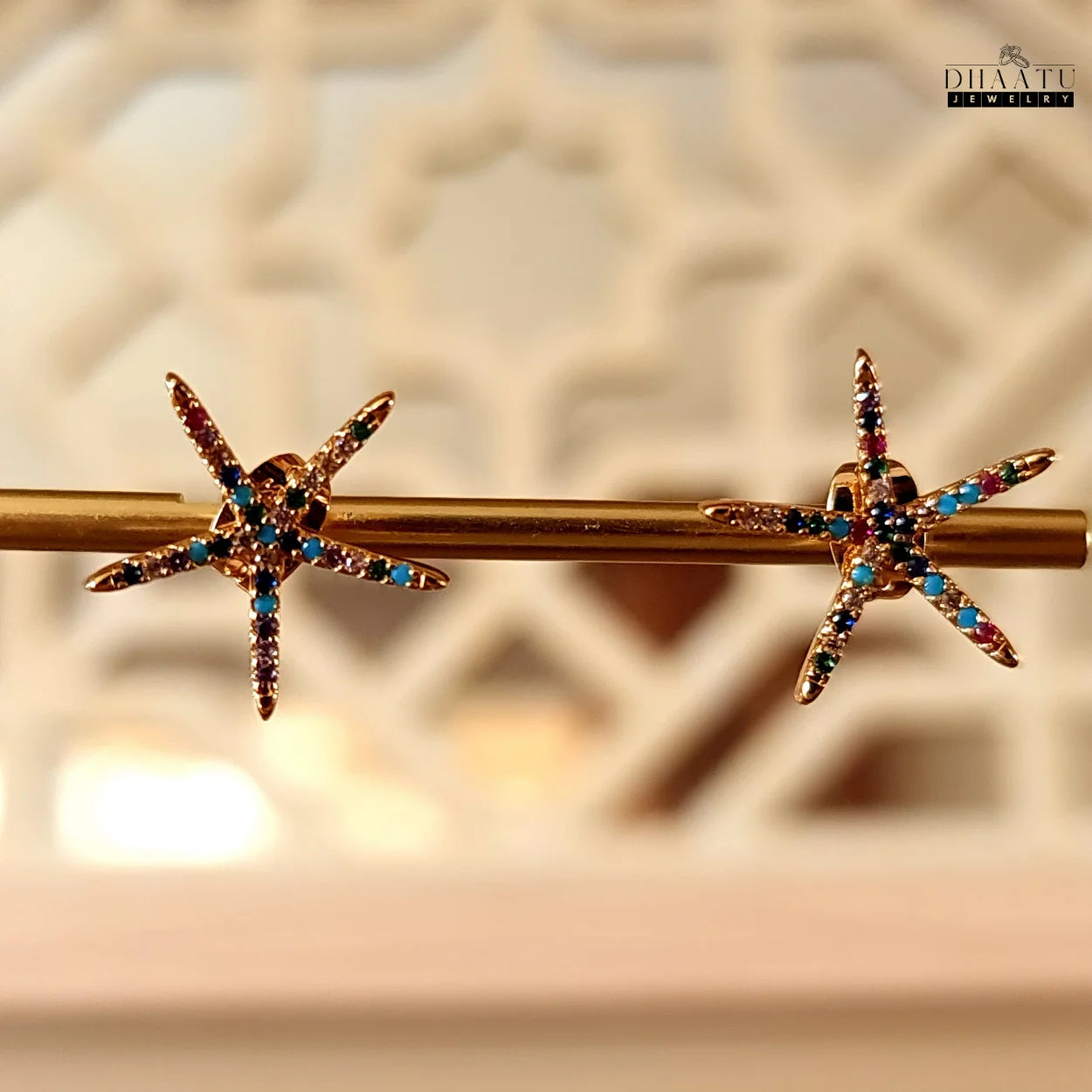 Vibrant rose gold starfish stud earrings with colorful zircon stones, elegantly displayed on a golden rod against a decorative, blurred background