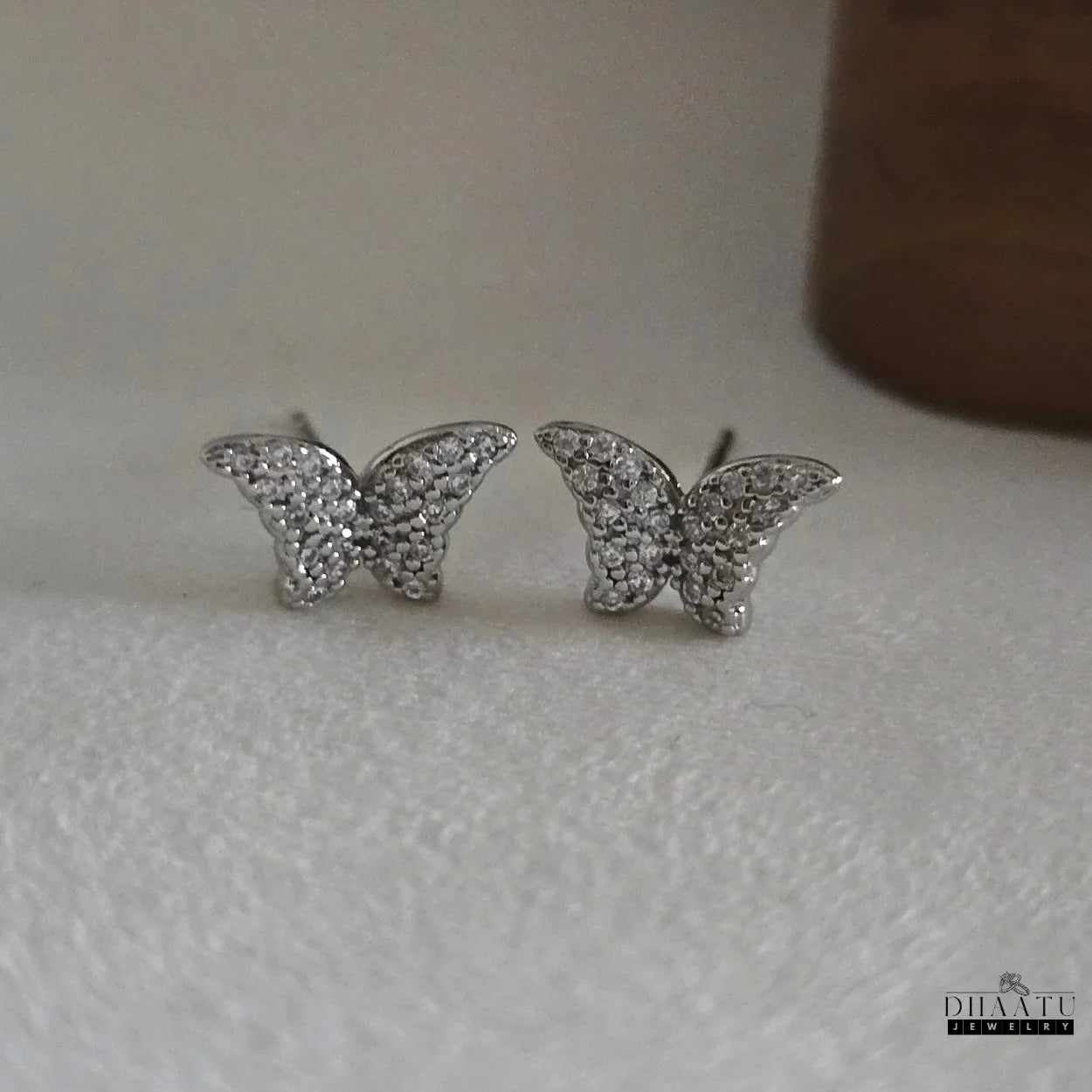 Front view of a pair of silver-toned butterfly stud earrings, adorned with brilliant zircon stones, on a white surface.

