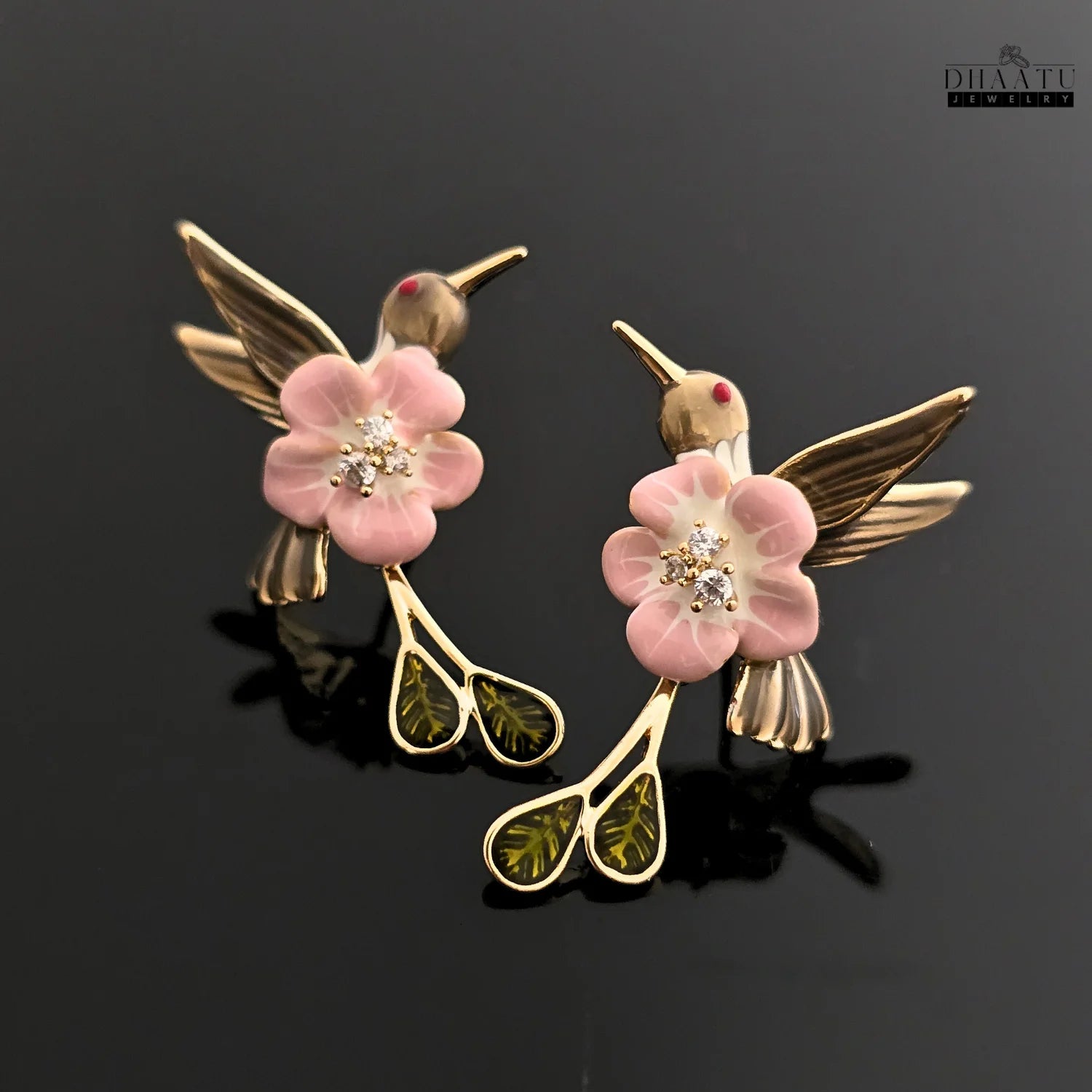 A pair of pink and brown enamel hummingbird and flower drop earrings on a black reflective surface.