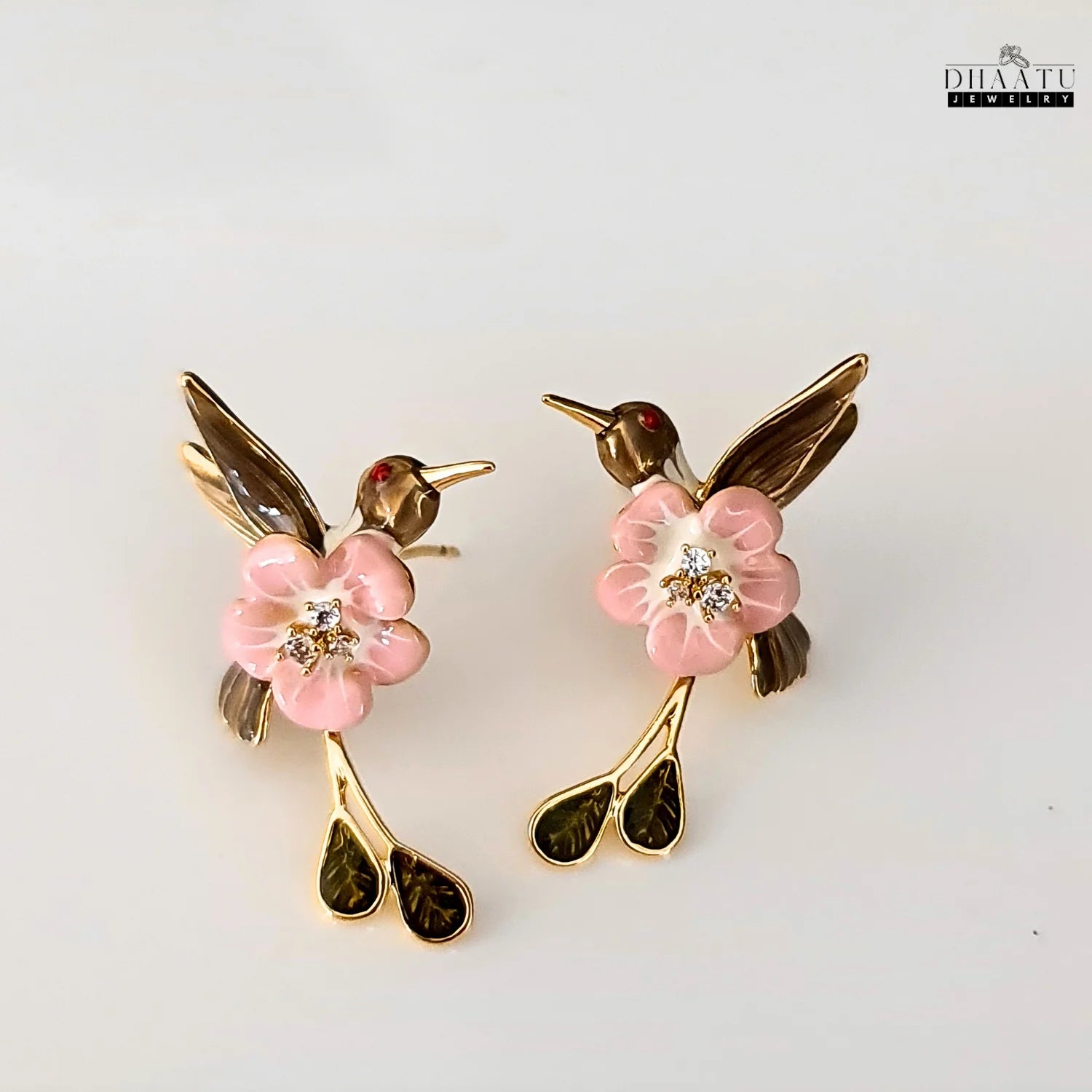 A pair of pink and brown enamel hummingbird earrings with a pink flower and leaves on a white background.