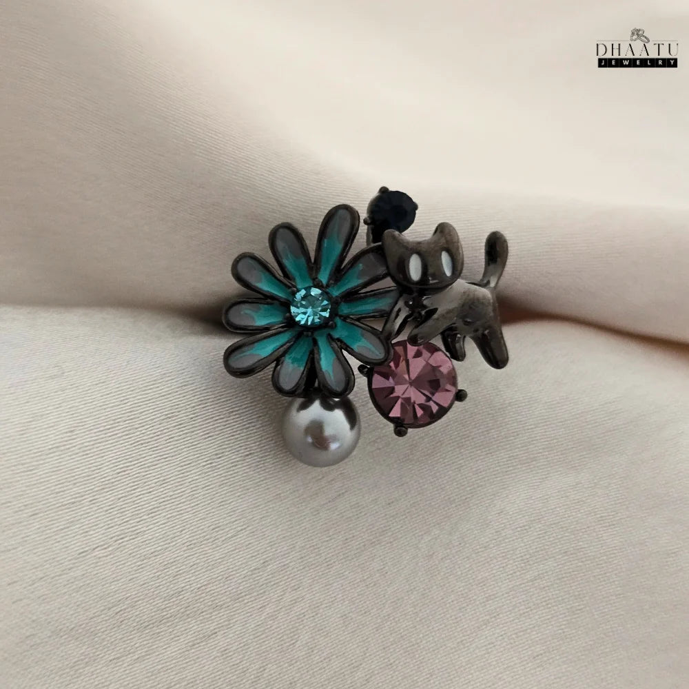 A close-up shot of the DHAATU JEWELRY playful cat and flower adjustable ring, showcasing the intricate teal enamel flower, pink rhinestone, and pearl drop on a neutral background.