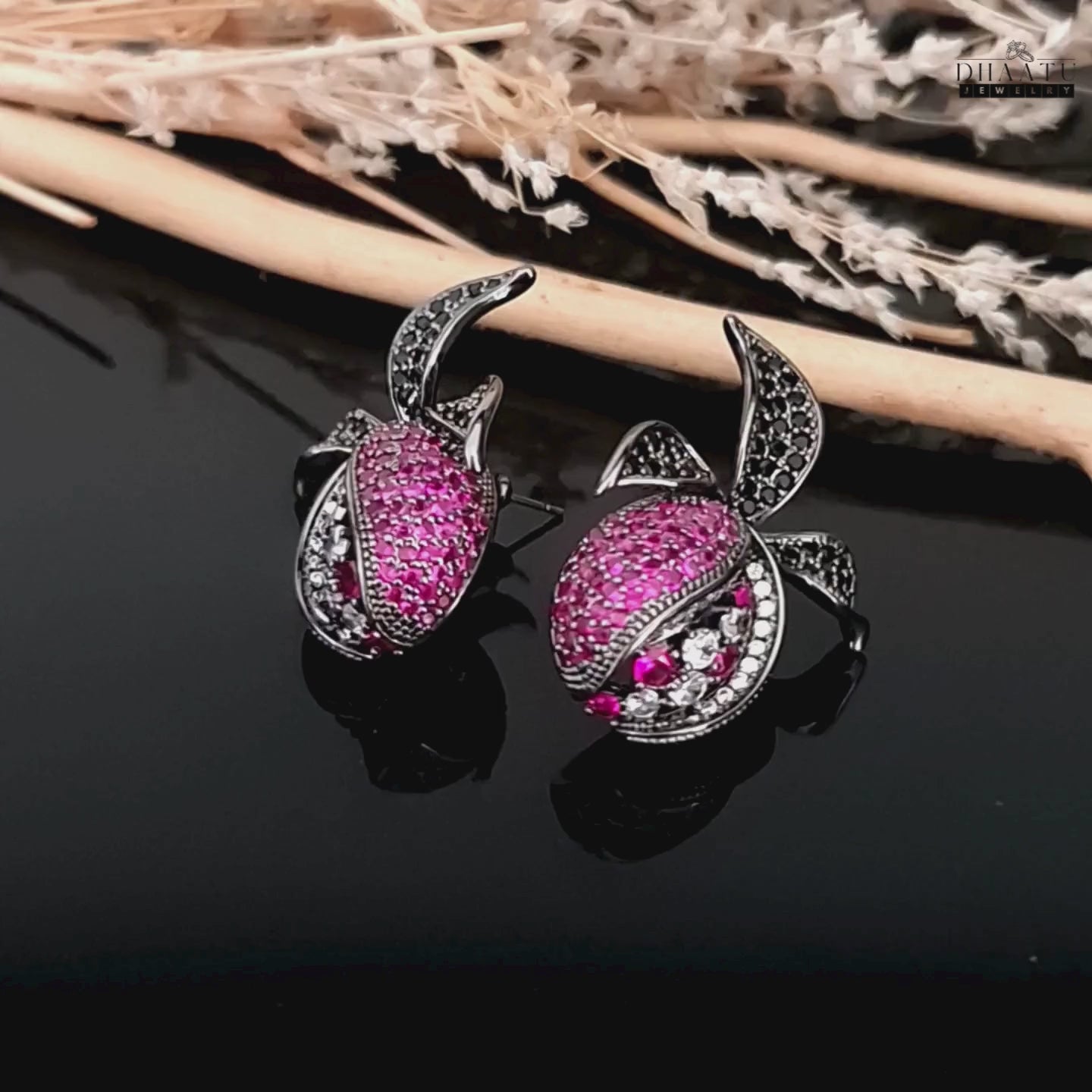 Load video: Product video of pomegranate-inspired stud earrings with black rhodium finish, featuring pink, white, and black zircon stones on a black reflective background.