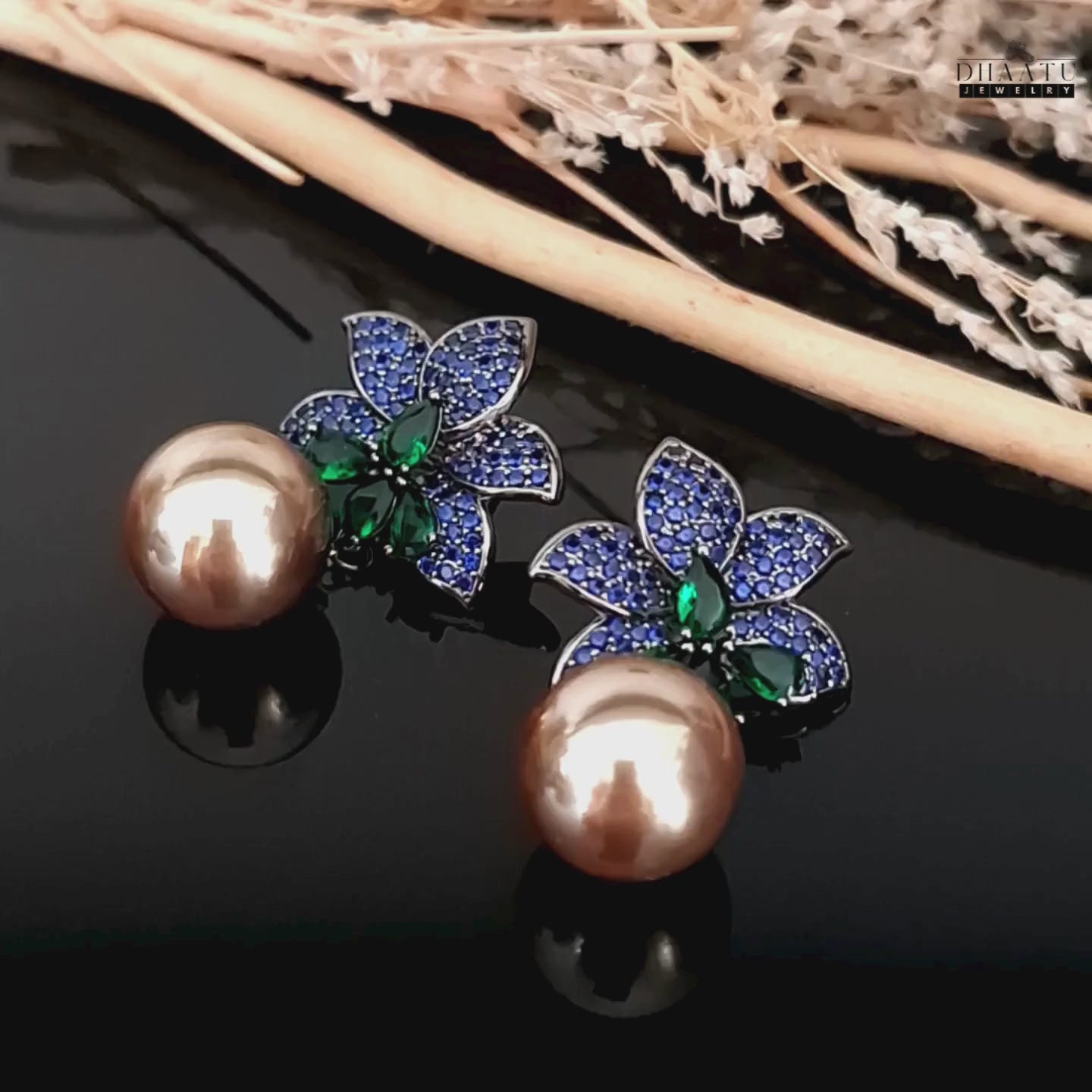 Load video: A video showcasing the Dhaatu Jewelry blue sapphire and emerald flower earrings with pearl drops, highlighting their intricate design and sparkle on a dark background with decorative elements.

