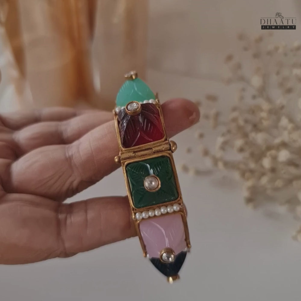 Load video: Video showcasing the multicolor carved stone and pearl kada bracelet being rotated by hand, highlighting its intricate details, hinged mechanism, and vibrant colors.
