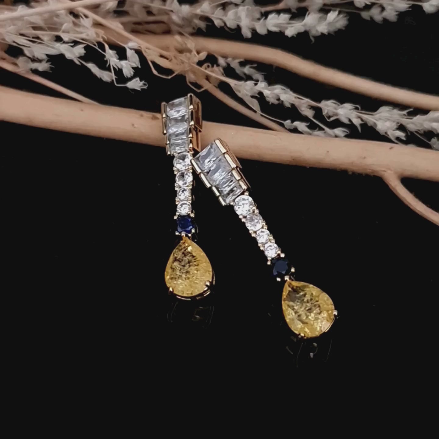 Load video: Product video of Gold-toned Zircon drop earrings with yellow and blue stones against a glossy black background
