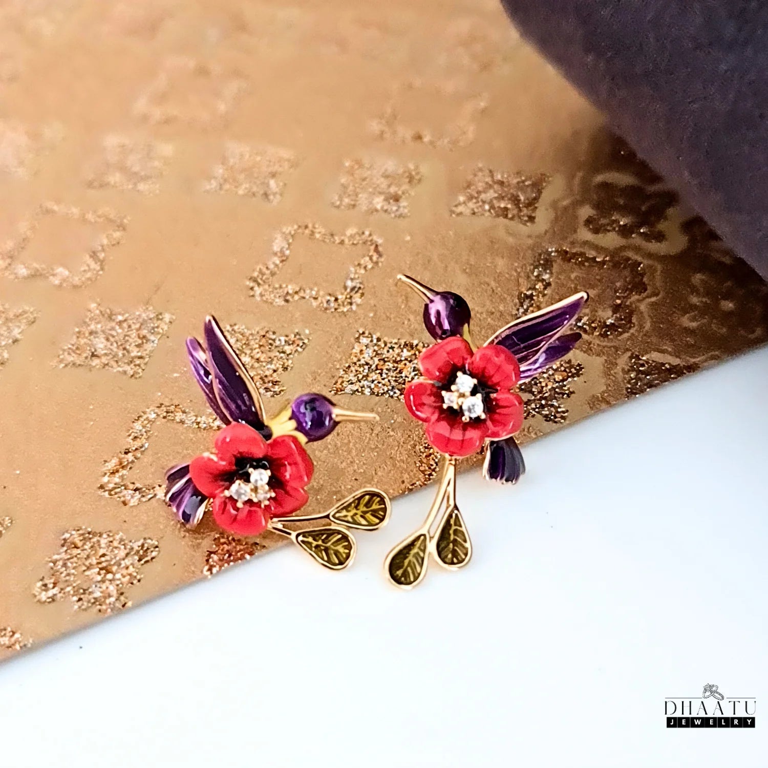 A pair of red and purple enamel hummingbird earrings styled on a textured gold and purple background.