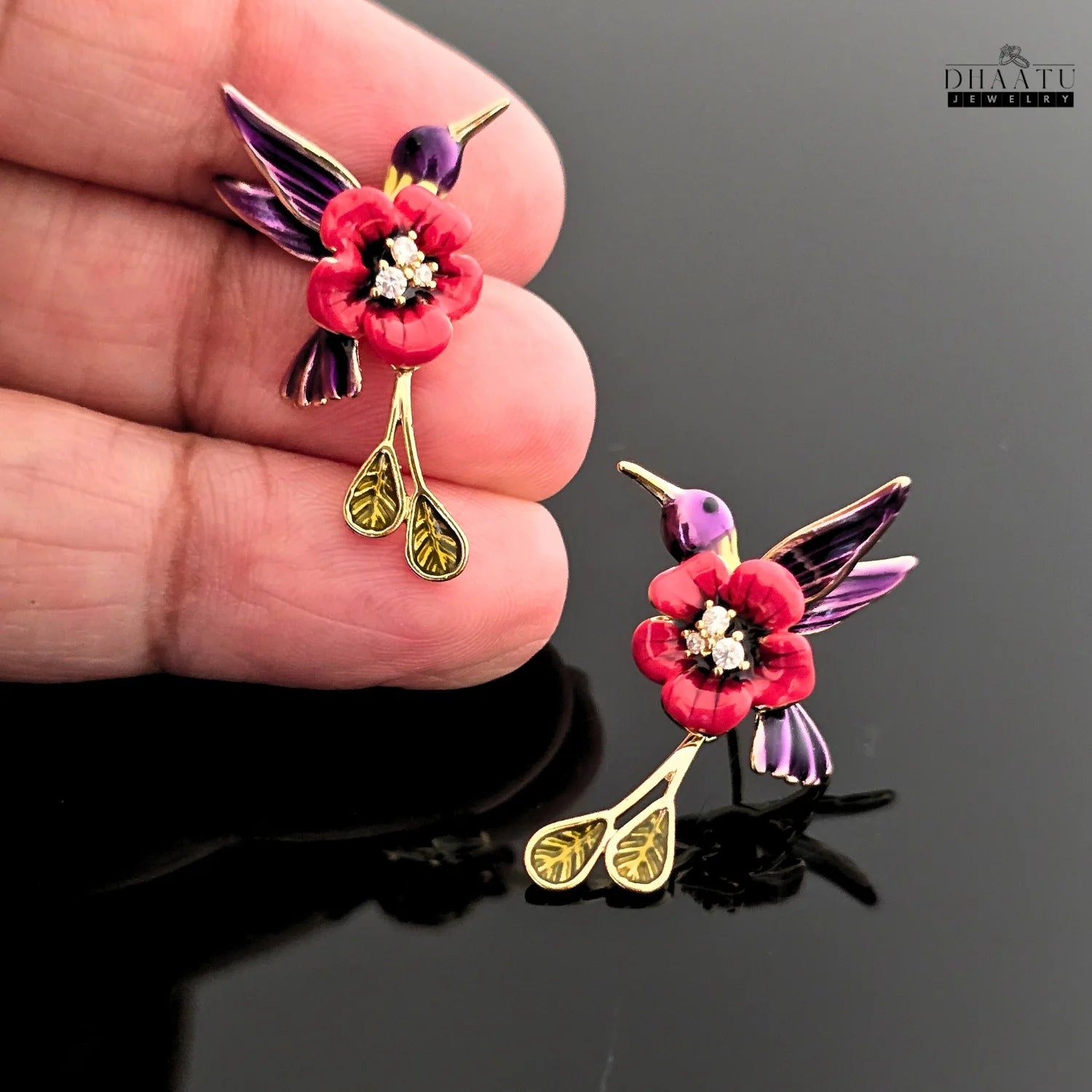 Close-up shot of a red and purple enamel hummingbird earring being held, showing intricate details and sparkling stones.