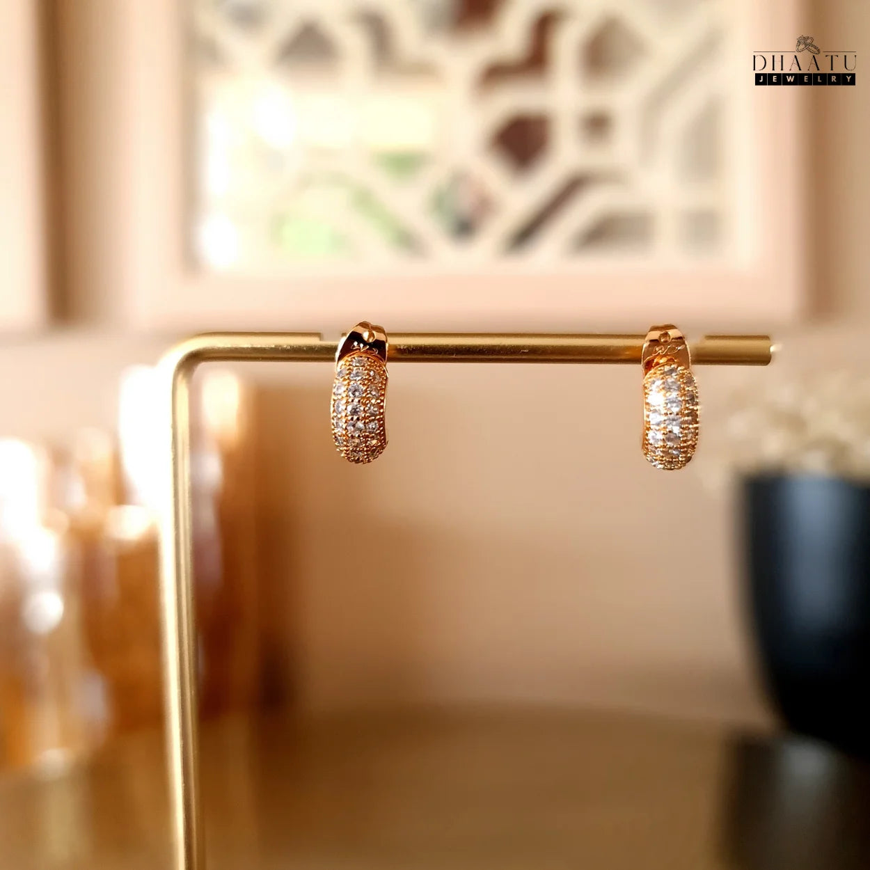 Rose gold plated huggie hoop earrings with zircon pave displayed on a golden jewelry stand against a blurred background, highlighting their elegant presentation.