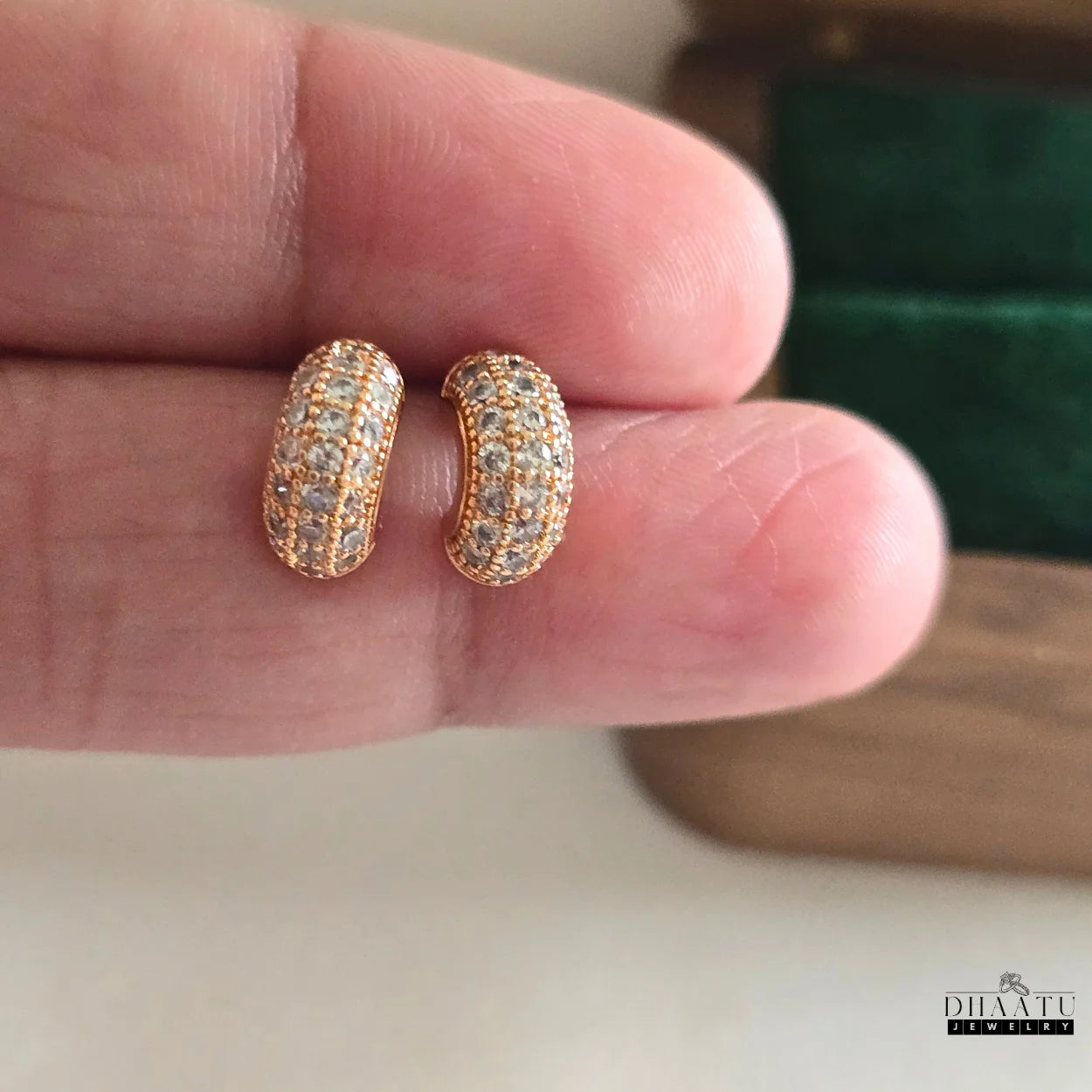 Two rose gold plated huggie hoop earrings with zircon pave held on fingertips, providing a sense of scale and detail of the jewelry.