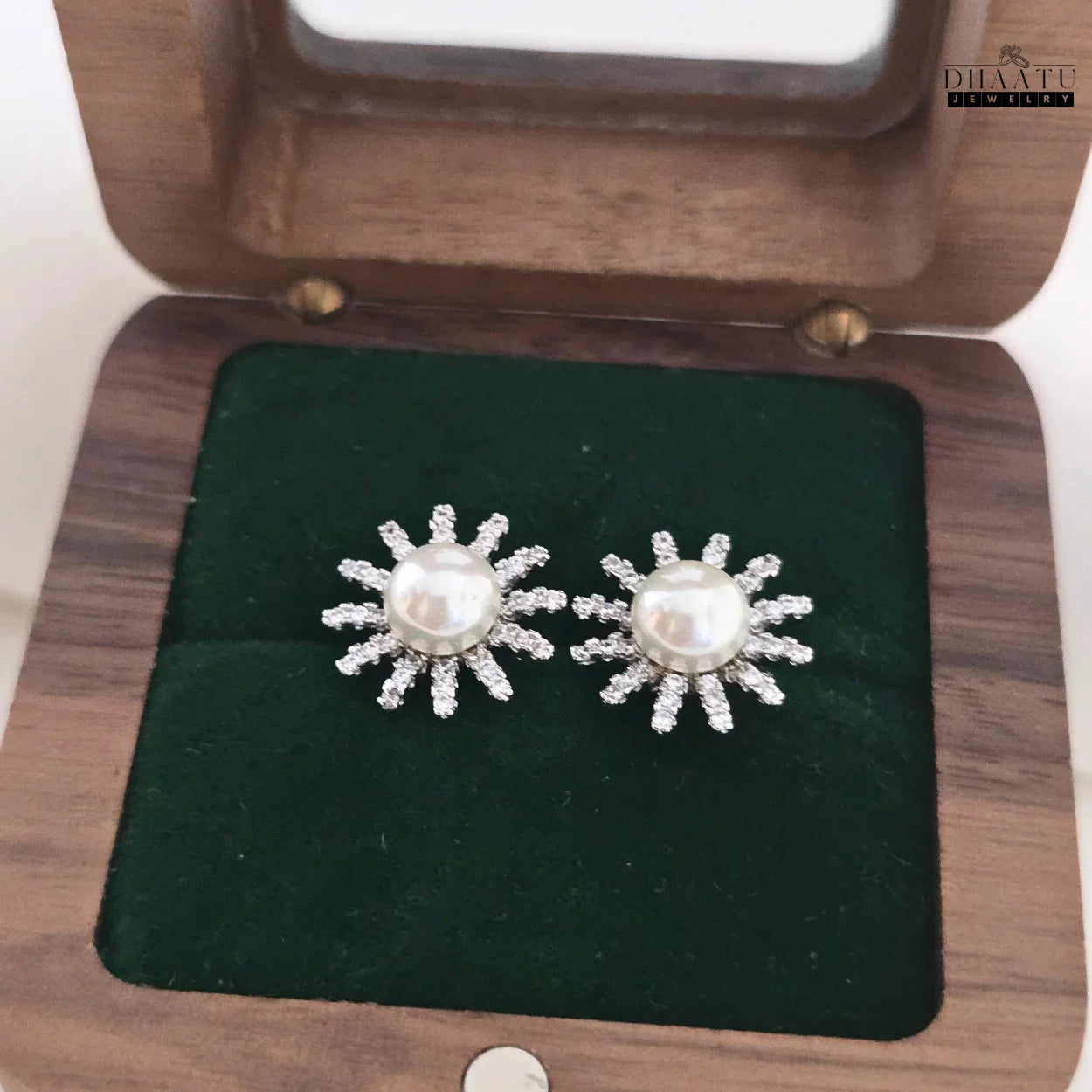 Two silver-plated sunburst stud earrings with white pearls and sparkling zircons resting on green velvet inside a wooden box.