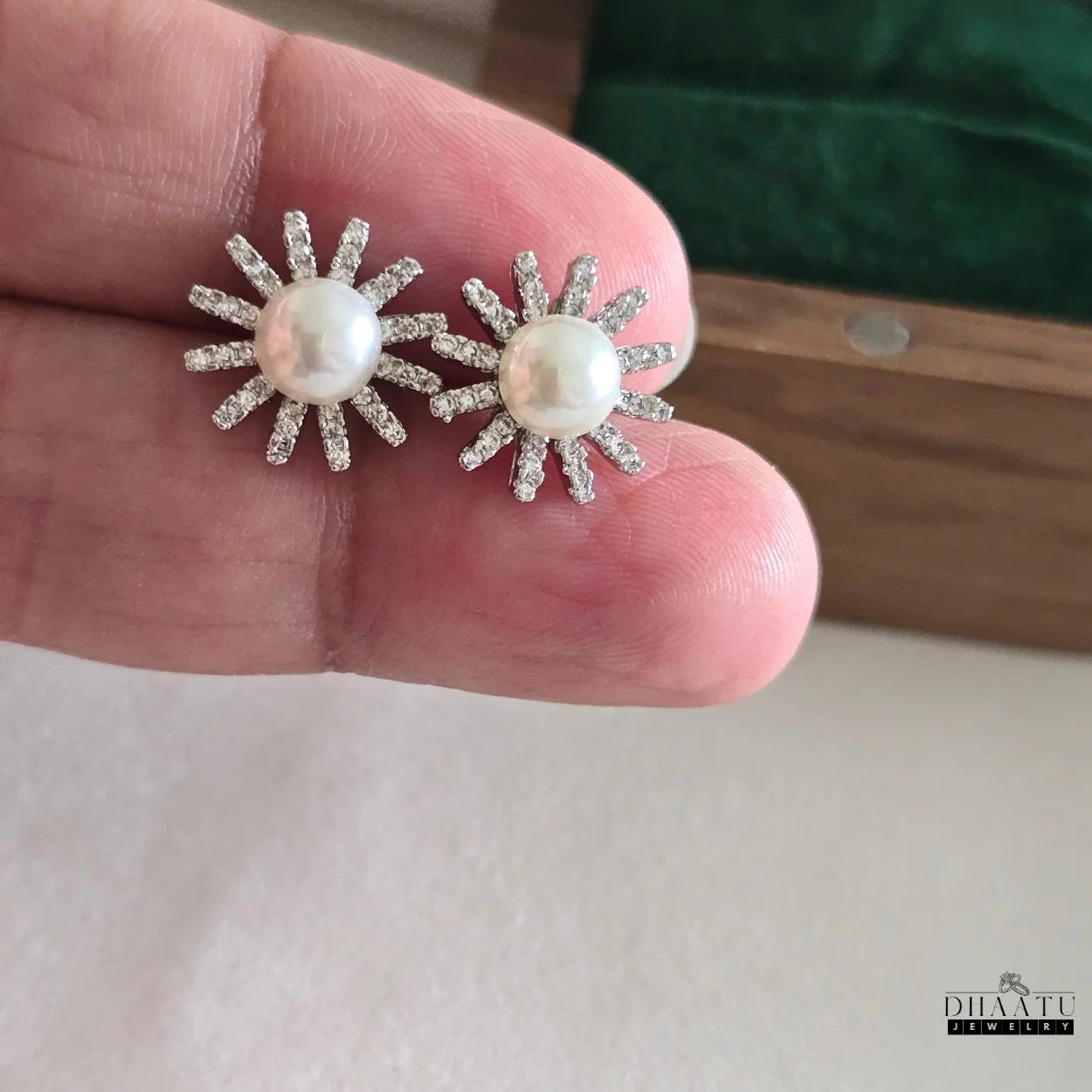 Close-up of two silver-plated sunburst stud earrings with pearls and zircons held on fingertips, showing size.

