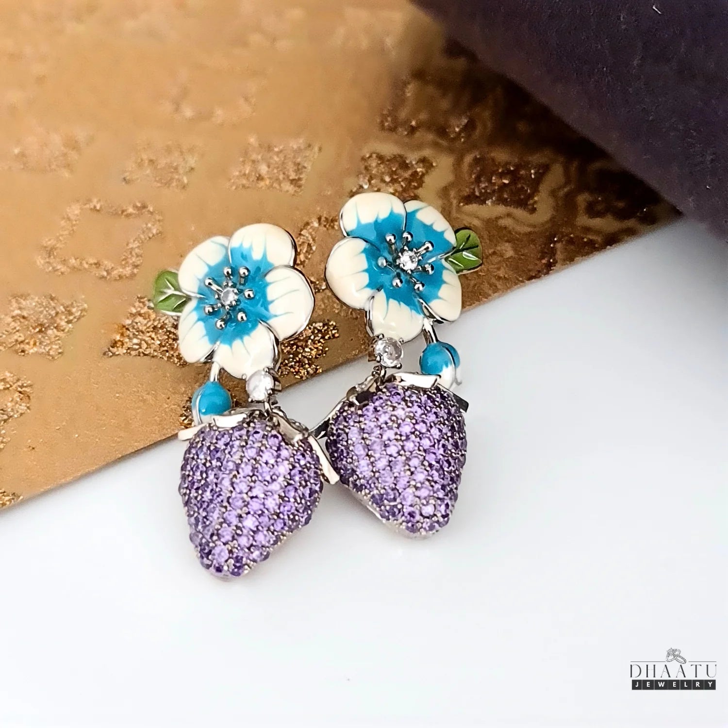 Silver-plated drop earrings with white and blue enamel flowers and purple CZ strawberry charms, displayed on a textured gold and white surface.