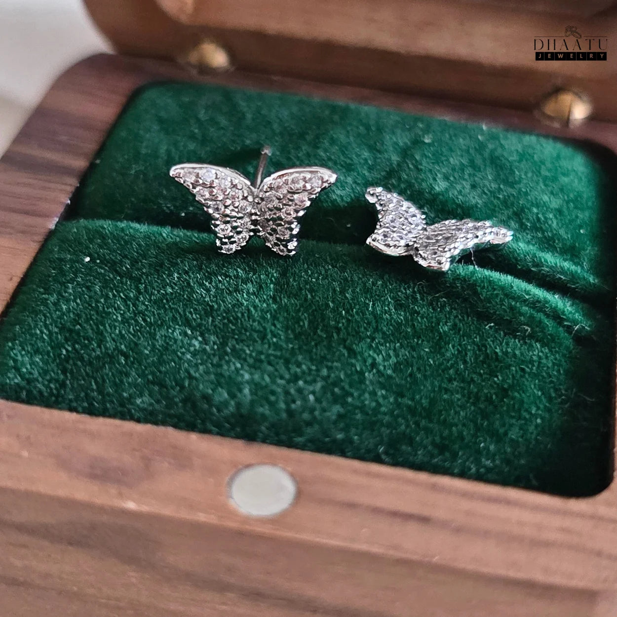 Close-up of sparkling silver-toned butterfly stud earrings with zircon stones, resting on green velvet inside a wooden jewelry box.


