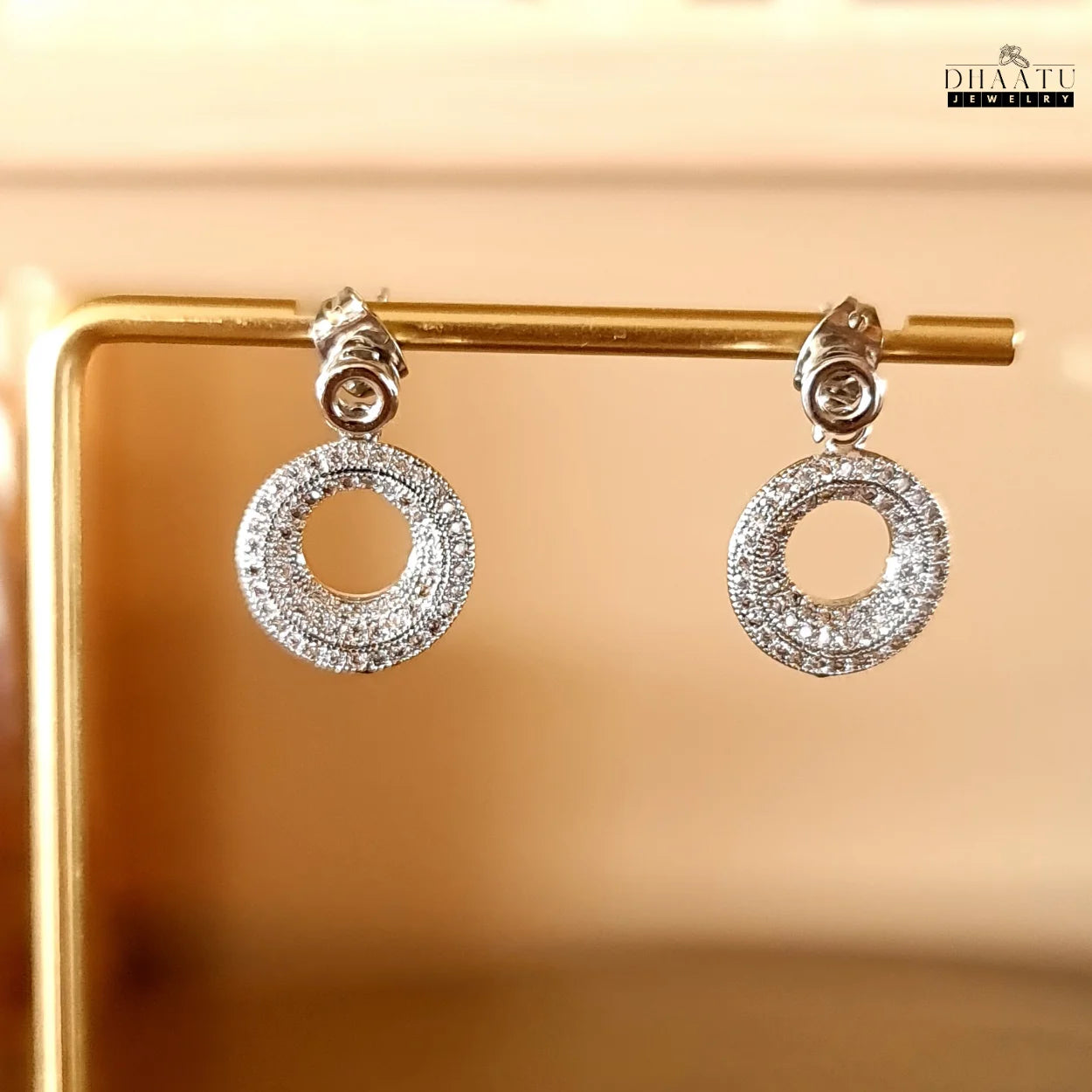 Close-up of sparkling circle stud drop earrings by Dhaatu Jewelry, showcasing pave set zircon stones on a golden display.