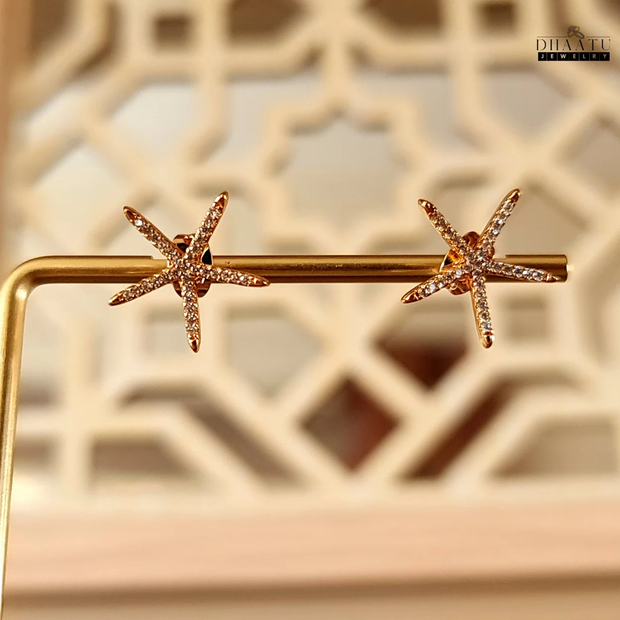 Sparkling rose gold starfish stud earrings with zircon stones showcased on a golden display stand against a blurred patterned background.

