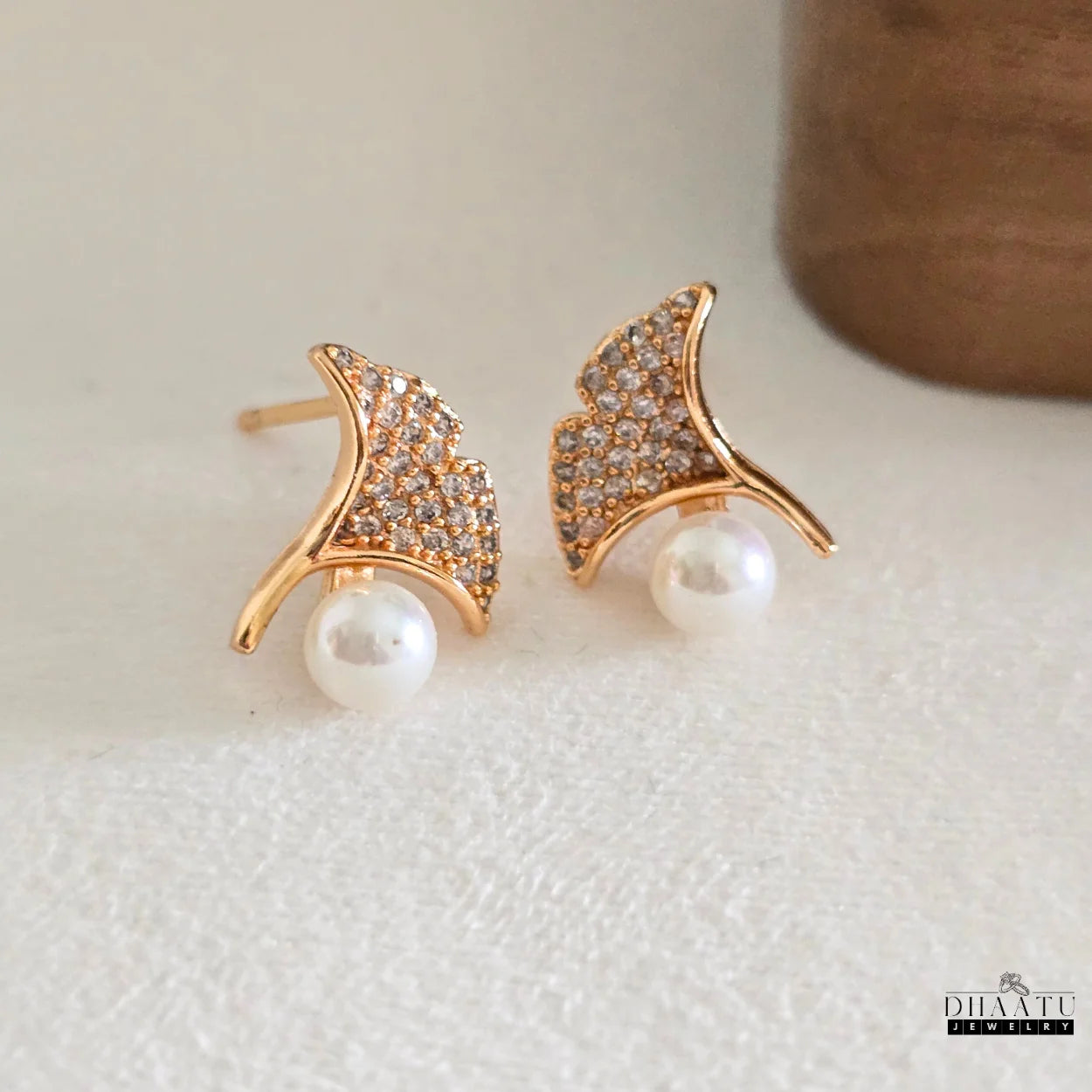 Stylish gold ginkgo leaf and pearl stud earrings with cubic zirconia, showcased on a textured white background.


