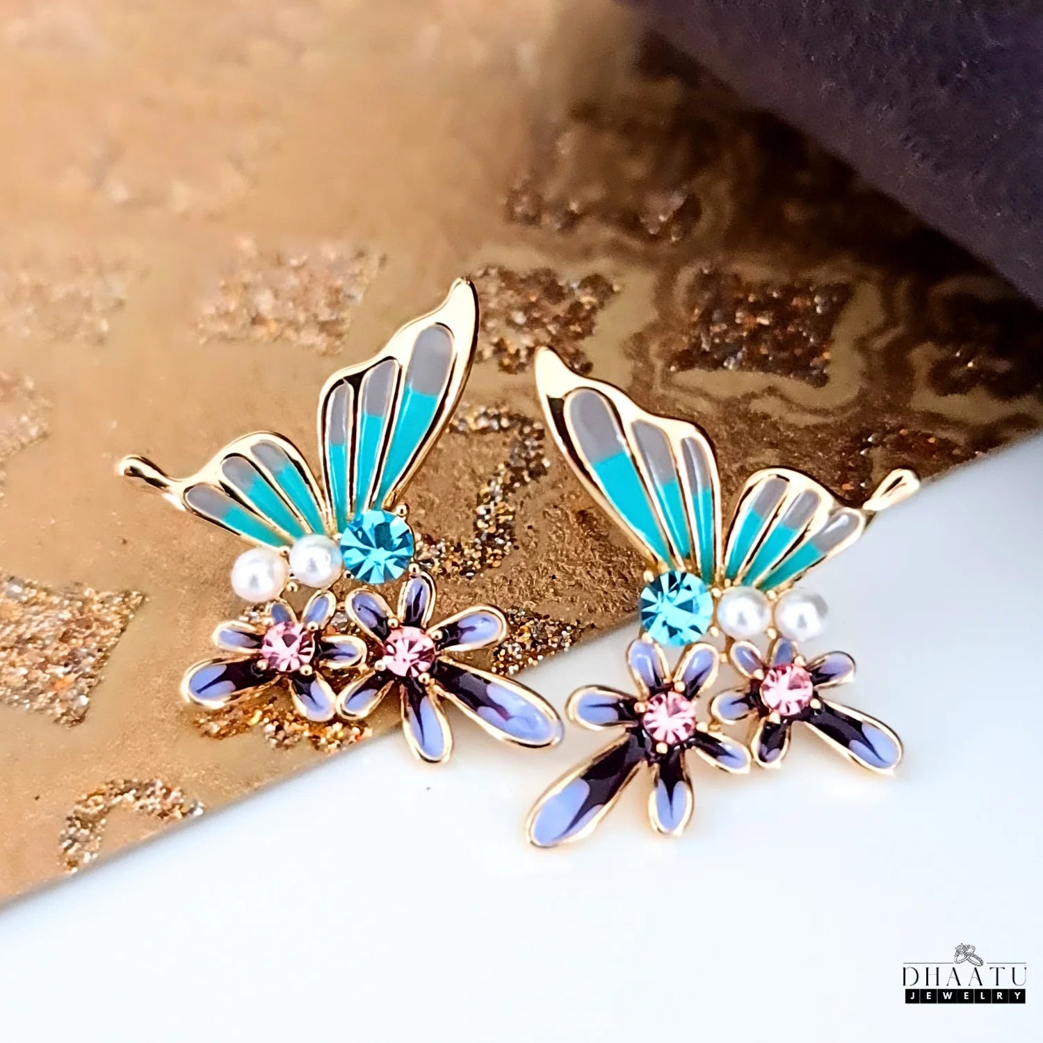 Pair of turquoise and purple butterfly and flower stud earrings on a gold textured background.