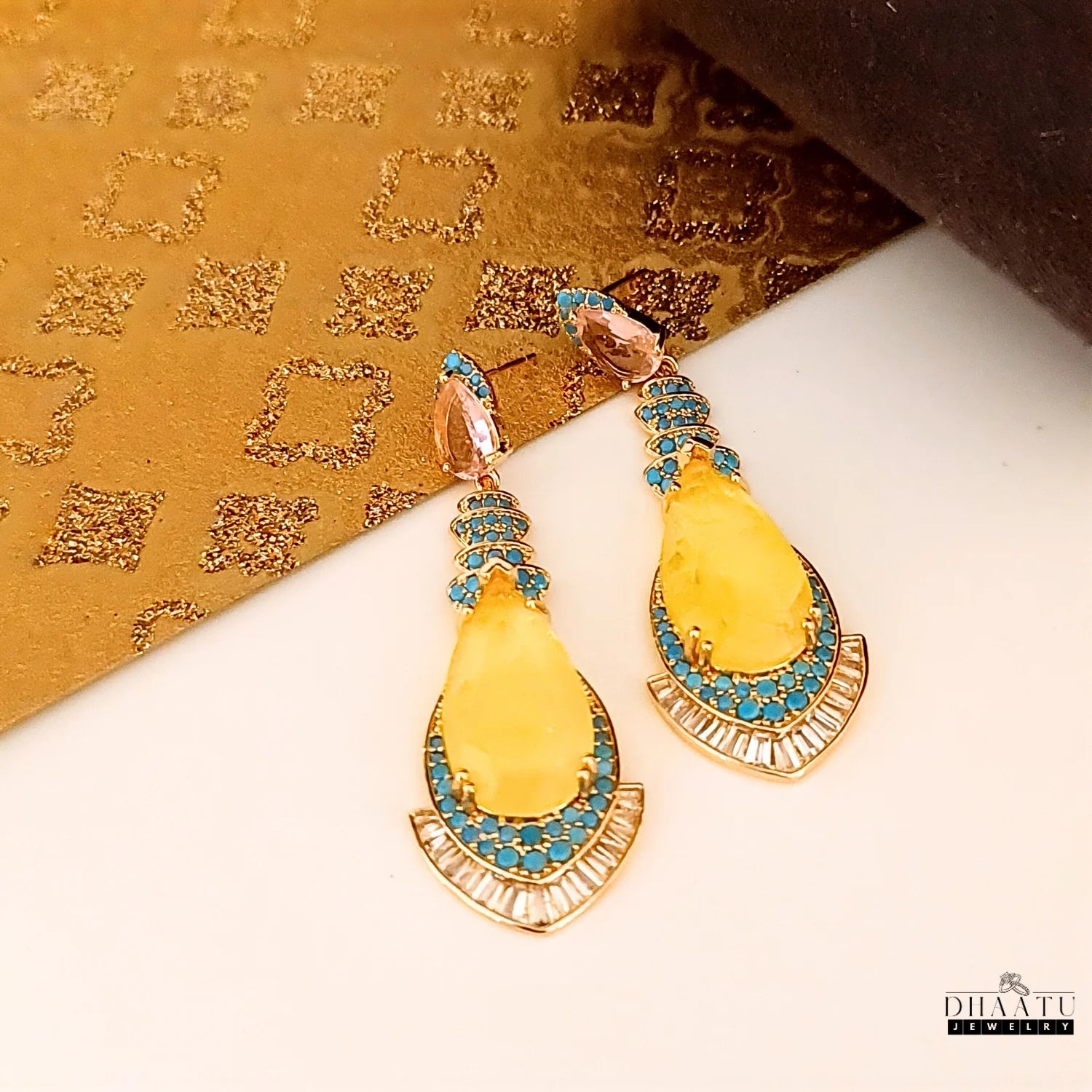 Close-up of artificial yellow doublet stone dangle earrings with turquoise and CZ on a luxurious gold textured background.